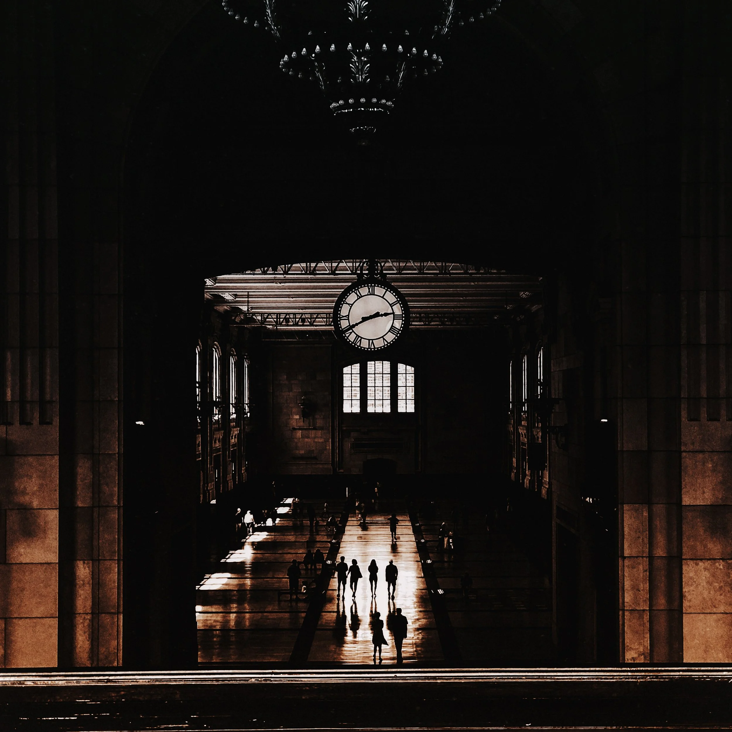 Silhouette of people walking inside a large, historic train station with a clock showing 5:40, high arched ceiling, and a chandelier hanging from it.