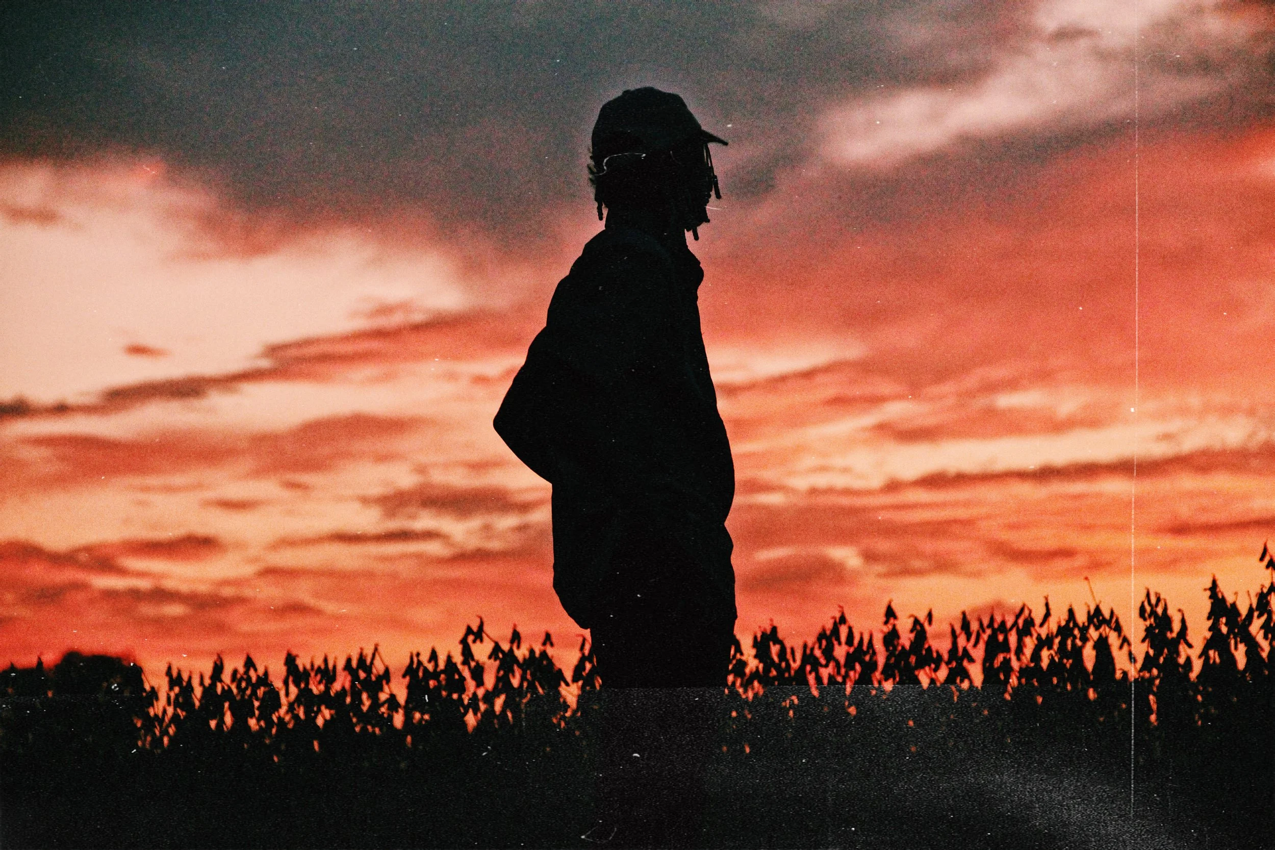 Silhouette of a person standing outdoors during a sunset or sunrise, with a colorful sky and trees in the foreground.