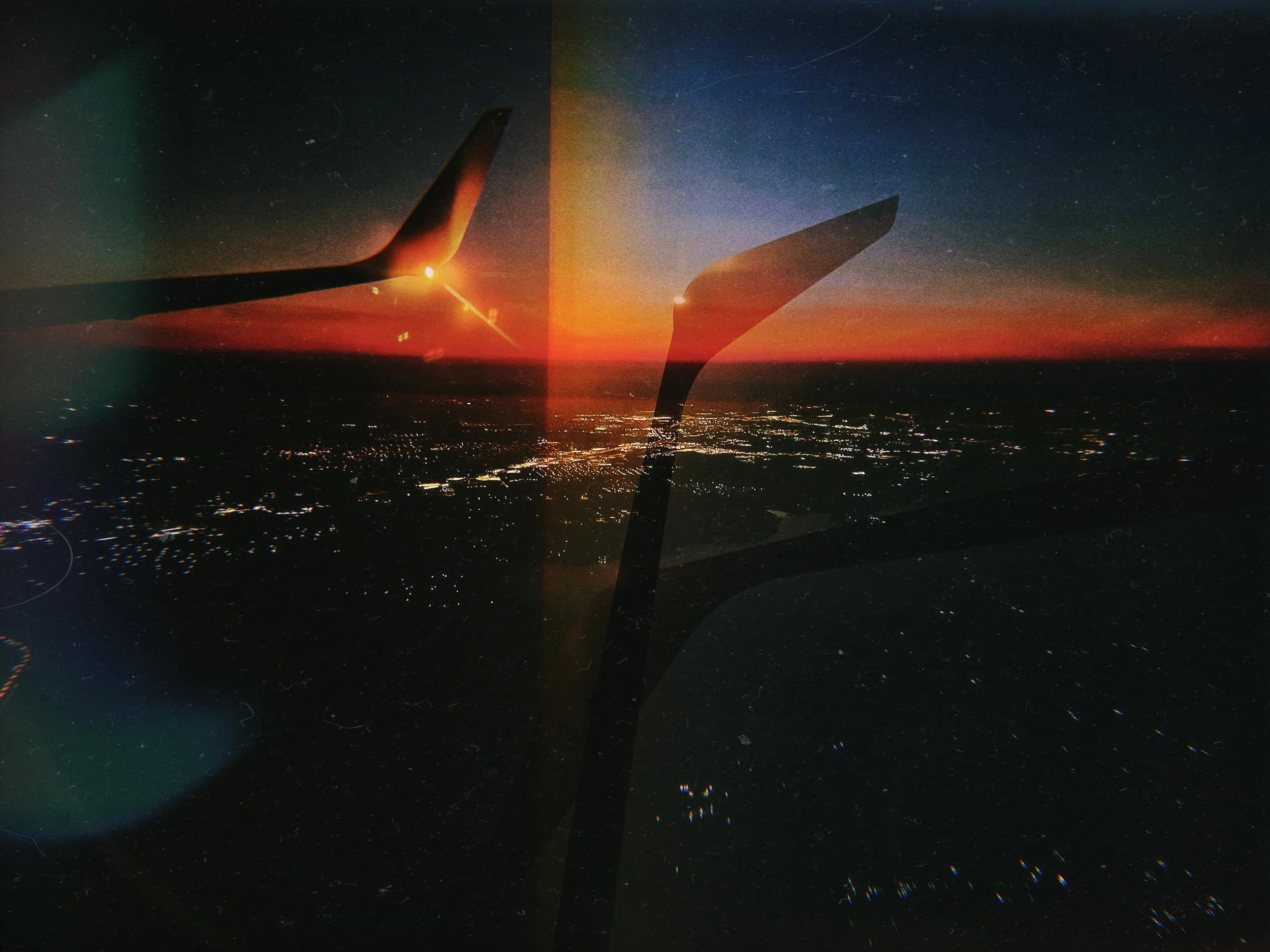 View from an airplane window showing the city lights below, a sunset with vibrant orange, pink, and purple colors, and a starry night sky with a faint rainbow-colored glow.