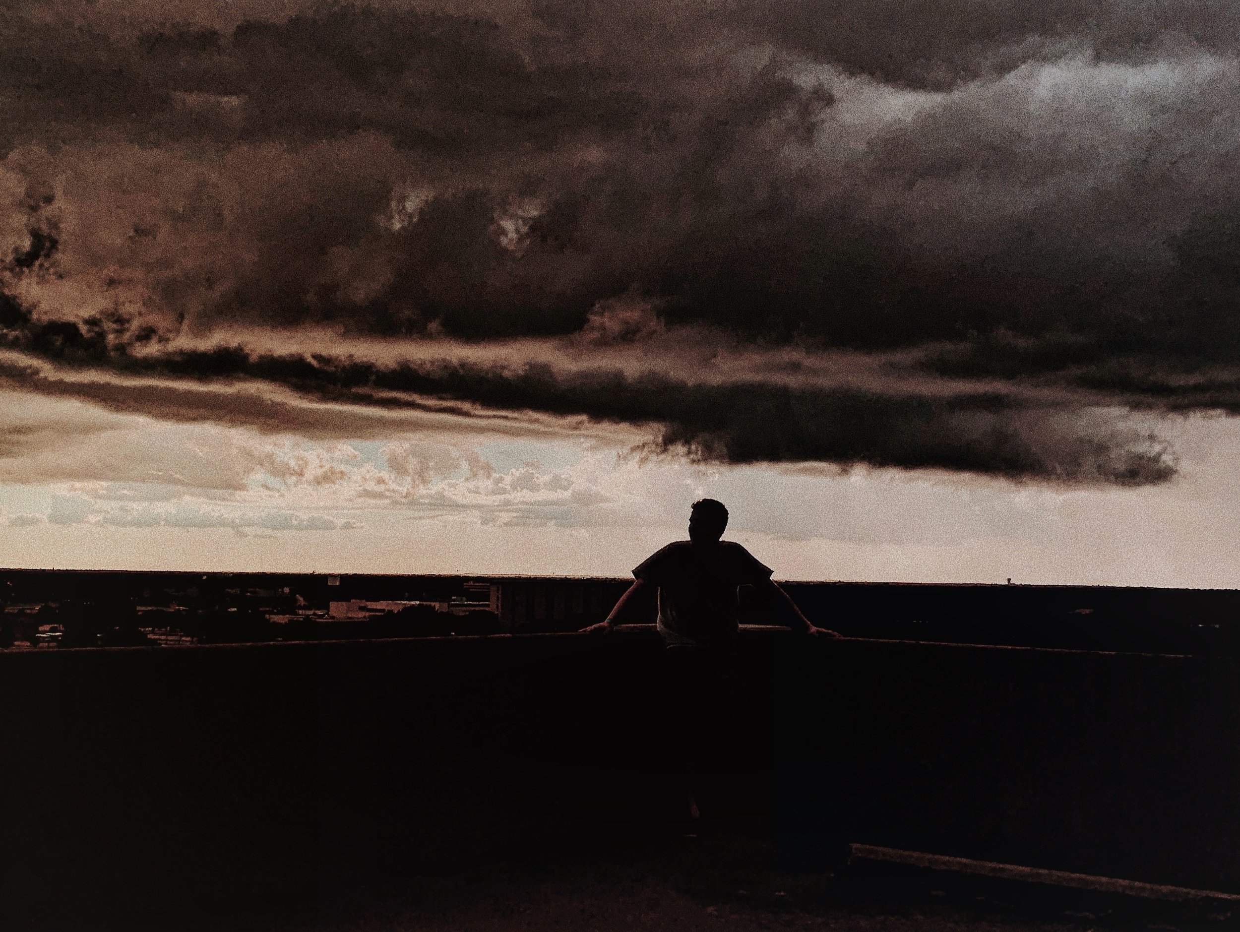 A person sits on a roof in front of a dark, stormy sky with thick, ominous clouds.