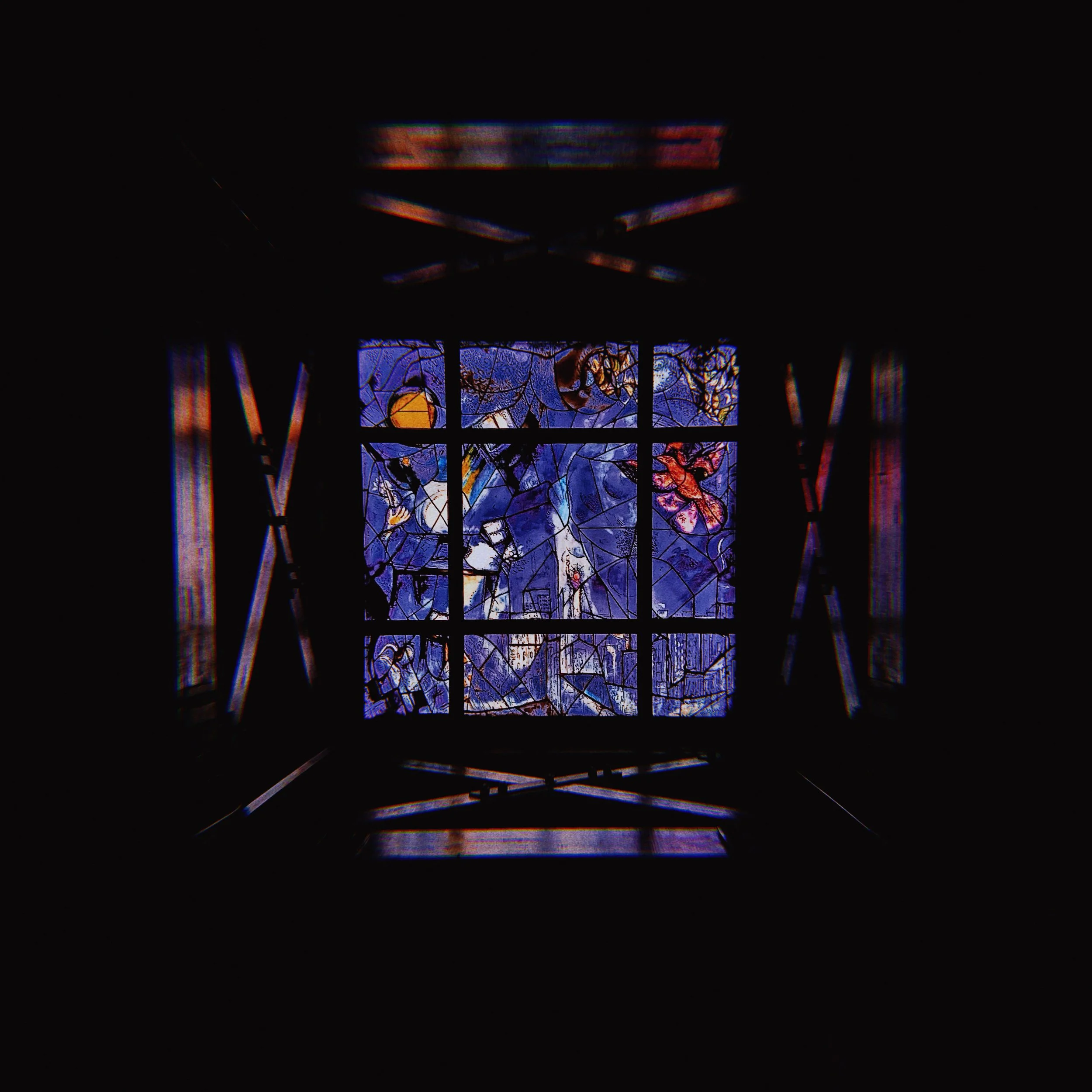 A stained glass window featuring colorful abstract designs with blue, orange, and purple hues, seen through a dark structure.