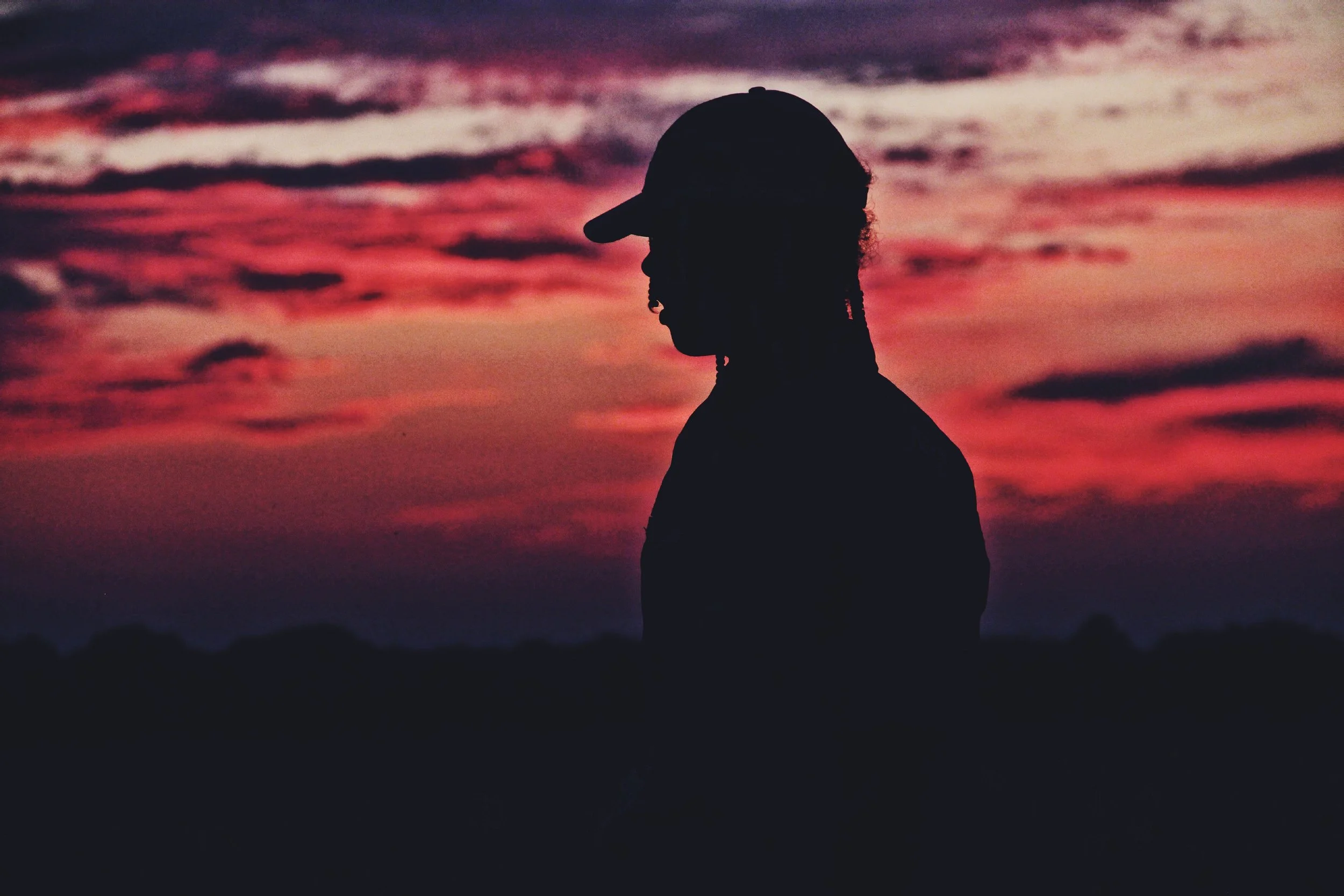 Silhouette of a person wearing a baseball cap, standing outdoors during sunset with a colorful sky filled with clouds.