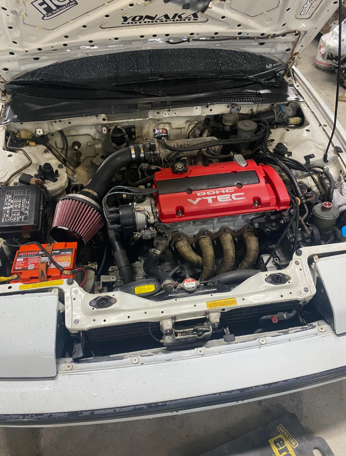 Close-up of a car engine bay featuring a red Honda DOHC VTEC engine cover, a cold air intake with a cone filter, and various engine components, with part of the car's white exterior visible.
