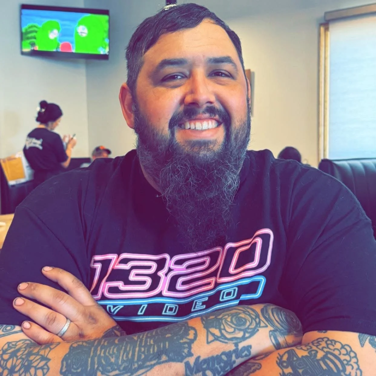 A smiling man with a beard and tattooed arms sitting at a table in a restaurant or cafe, wearing a black t-shirt with neon text. In the background, a woman is standing and looking at her phone, and a TV screen is mounted on the wall.