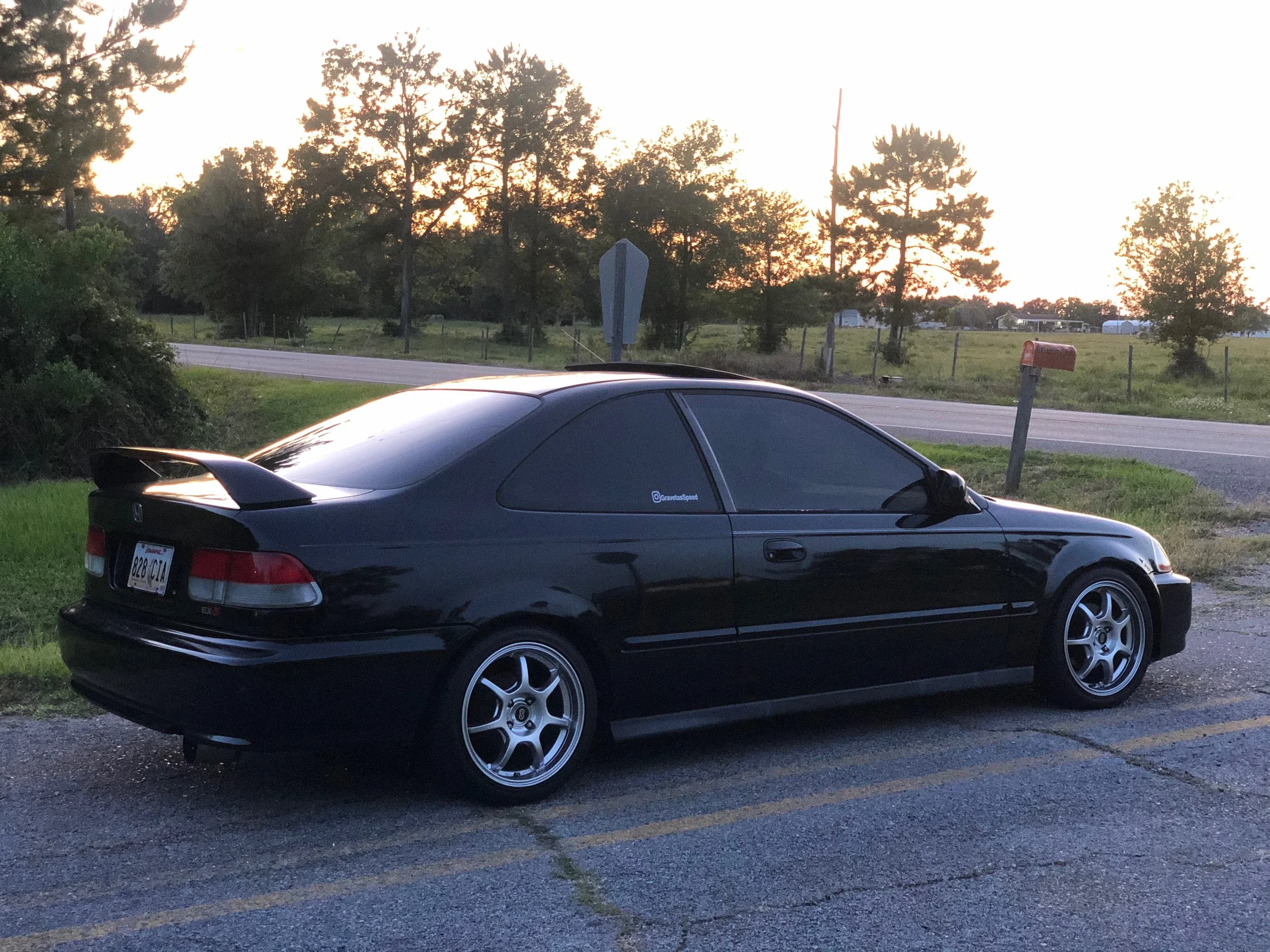 A black sports car parked on the grass beside a rural road at sunset, with trees, a mailbox, and a fence in the background.