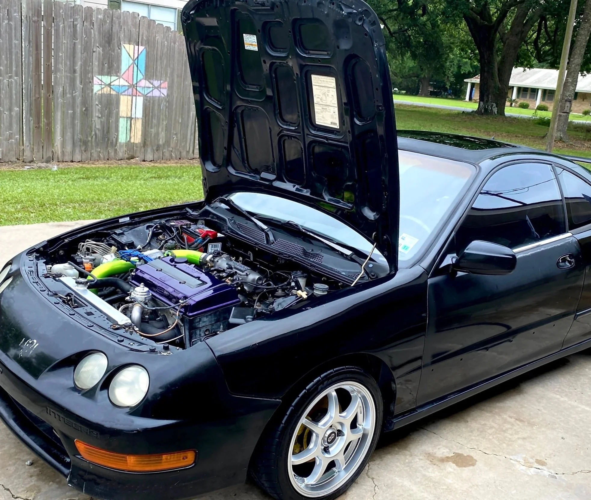 A black sports car with its hood open, showing the engine inside, parked outdoors in a residential area with trees, a wooden fence with a colorful geometric design, and a house in the background.