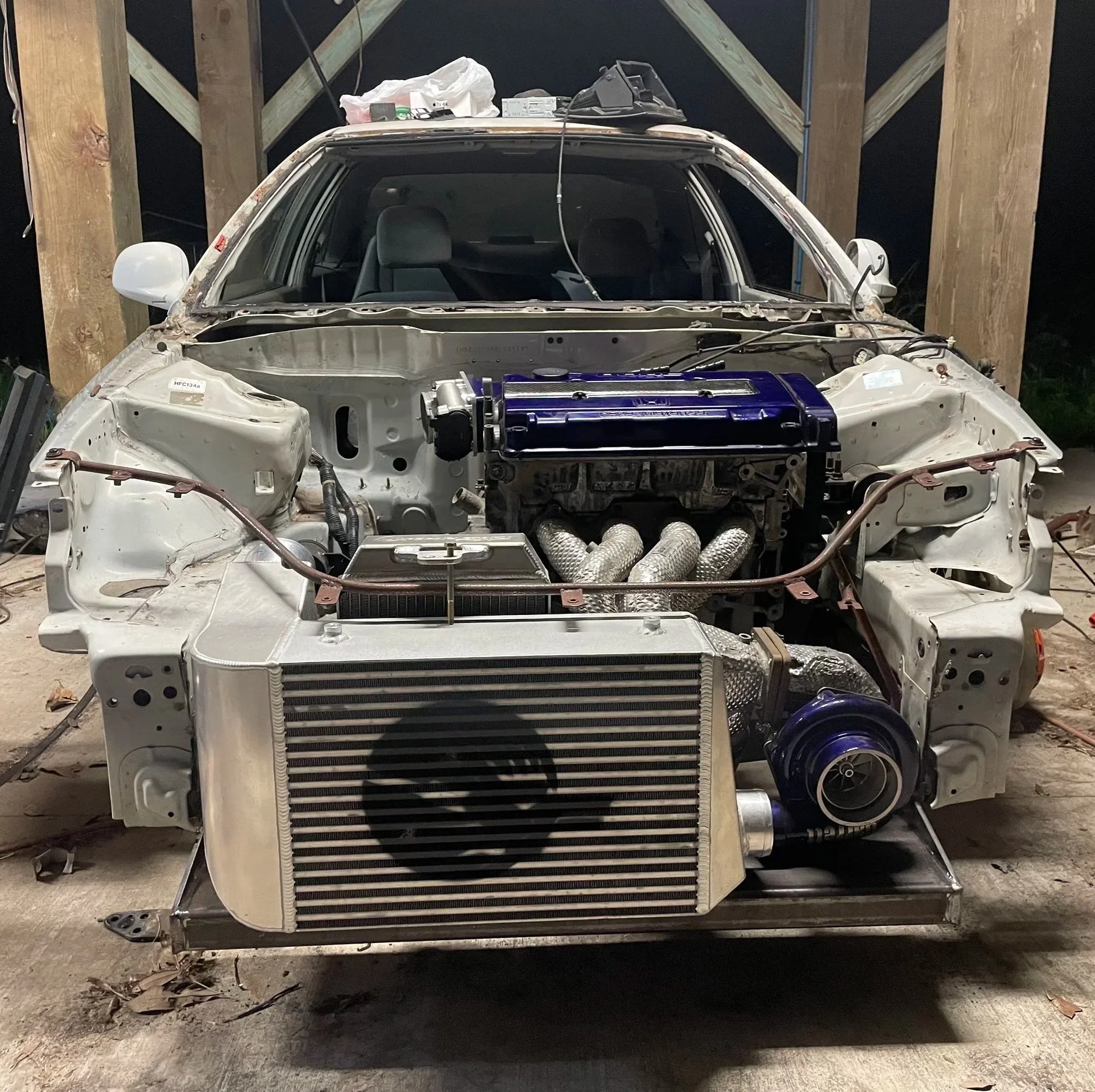 The image shows a car under restoration with its bodywork removed, revealing the engine, radiator, and exhaust system. The surrounding area appears to be a workshop or garage with wooden beams and tools on top of the car.