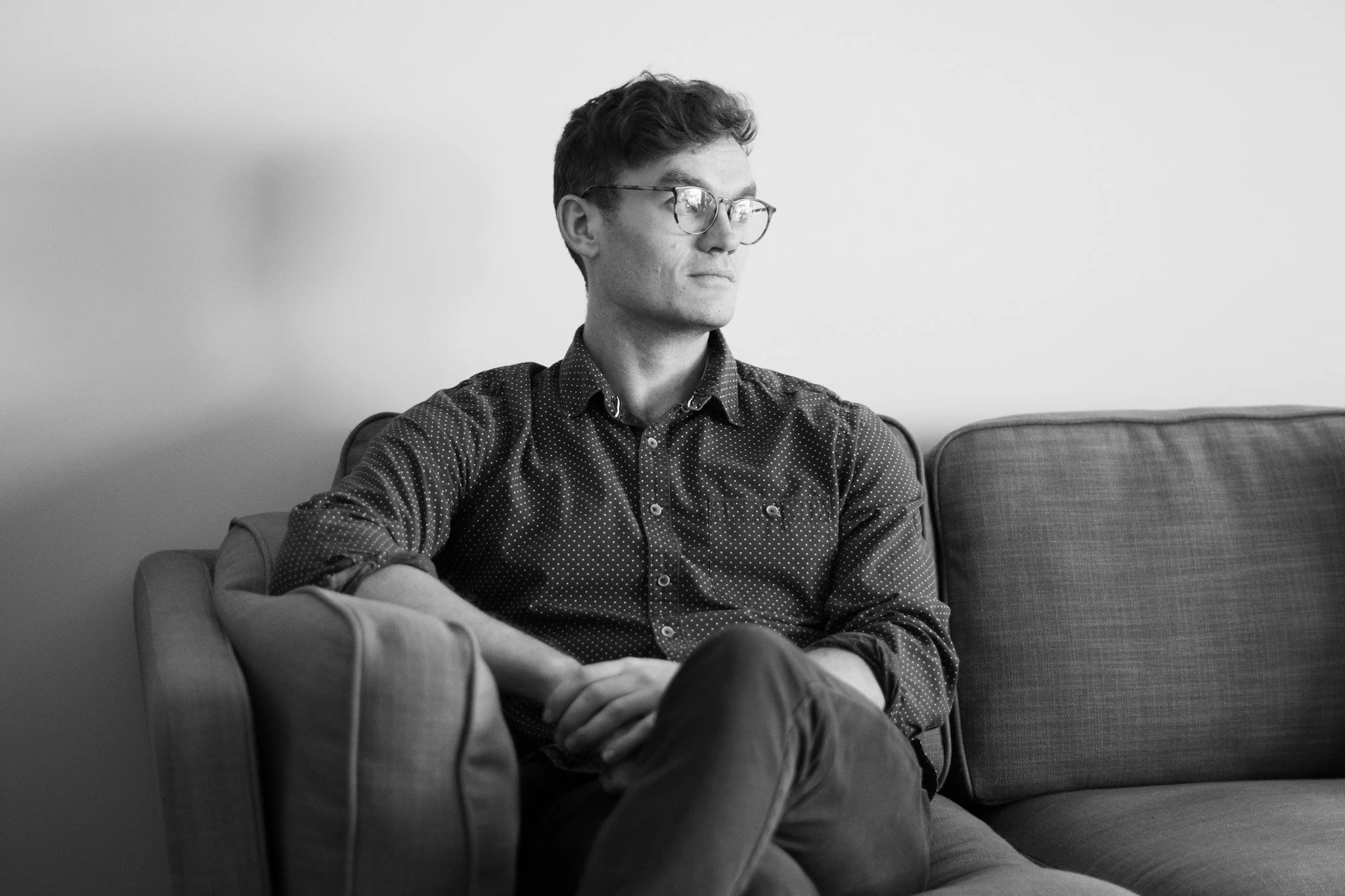 A young man with glasses sitting on a couch against a plain wall, wearing a patterned shirt with sleeves rolled up, looking to the side with a serious expression.