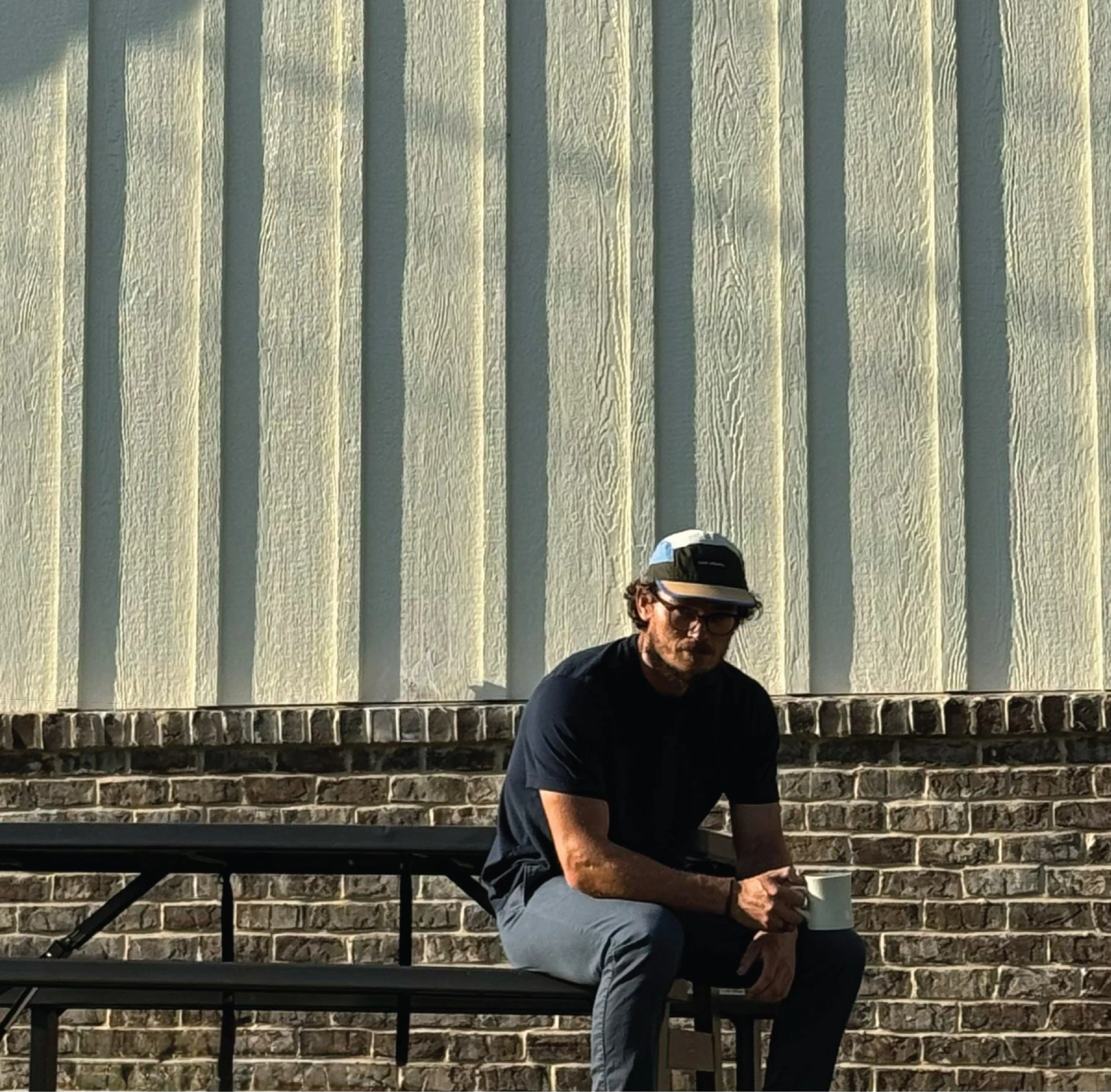 A man with dark hair, glasses, and a beard, wearing a black t-shirt, sitting on steps in front of a brick wall.