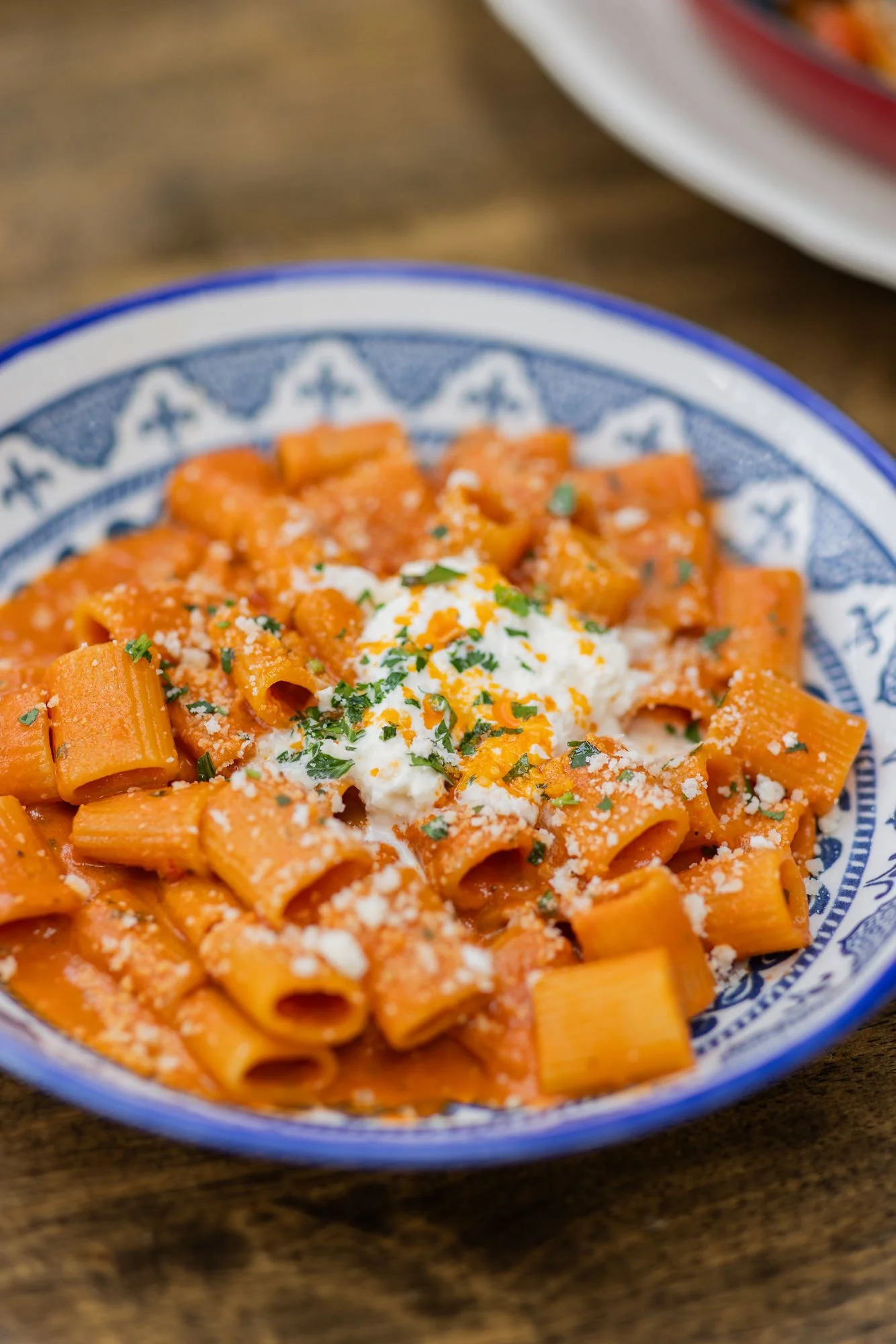 Close-up of a bowl of baked ziti pasta with tomato sauce, topped with fresh herbs and grated cheese on a wooden surface.