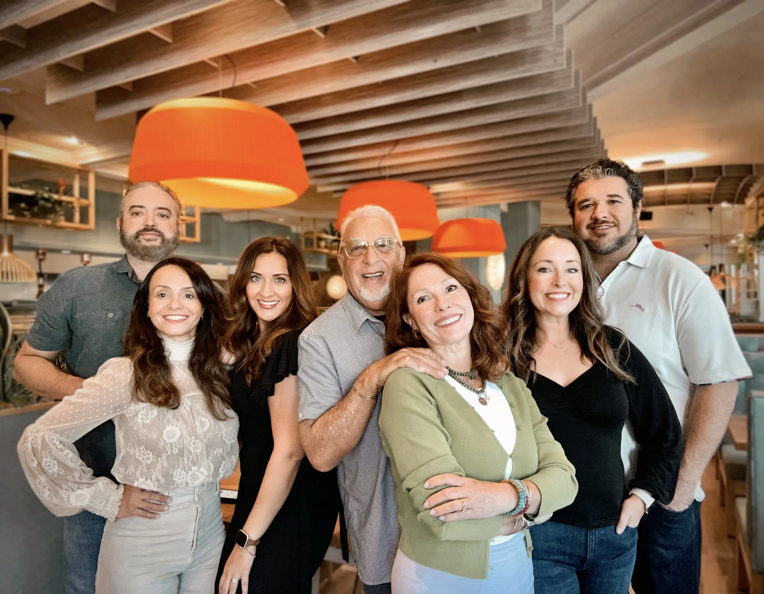 A group of nine diverse people smiling and posing together in a warmly lit restaurant or café with orange pendant lights and wooden ceiling beams.