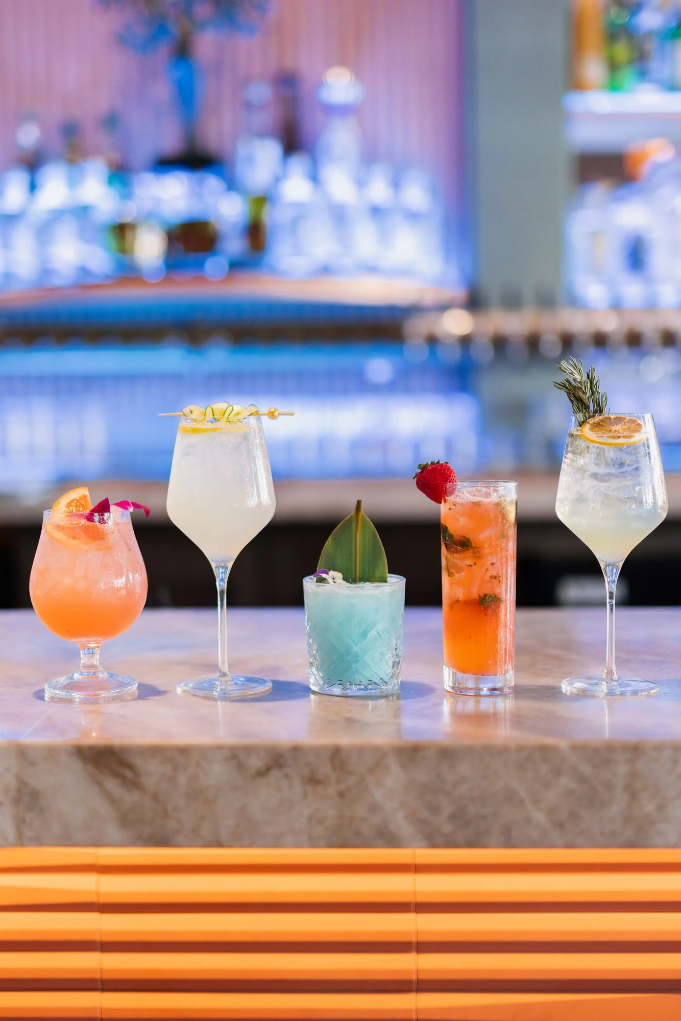 A variety of colorful cocktails on a bar counter in front of blurred bottles and bar equipment.