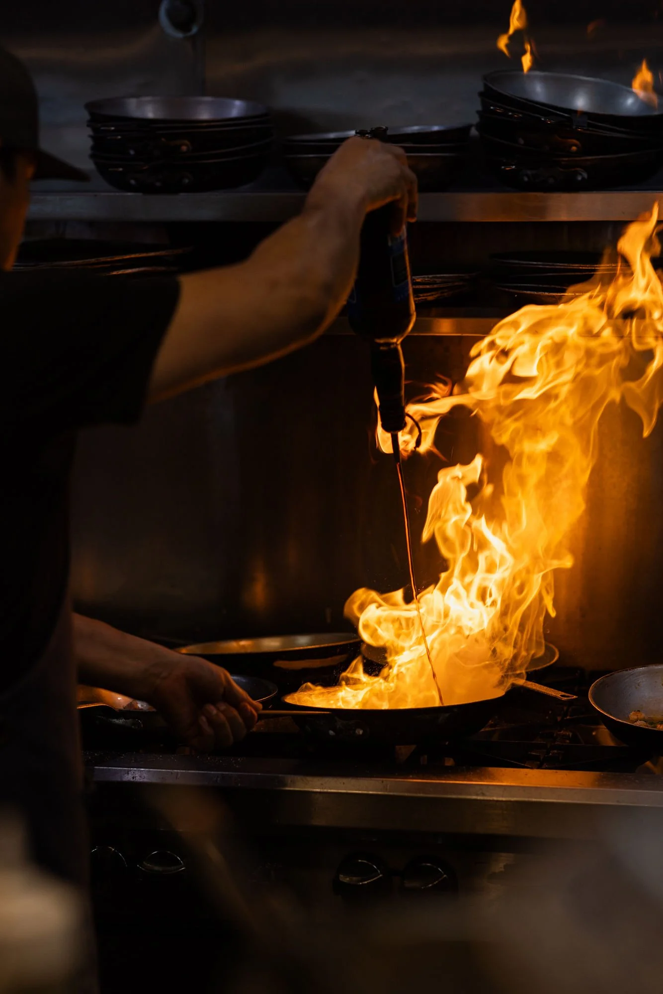 A chef is cooking with a flame in a pan on a commercial stove. The chef’s arm and hand hold a bottle, possibly adding an ingredient, while the stove is actively producing bright flames.