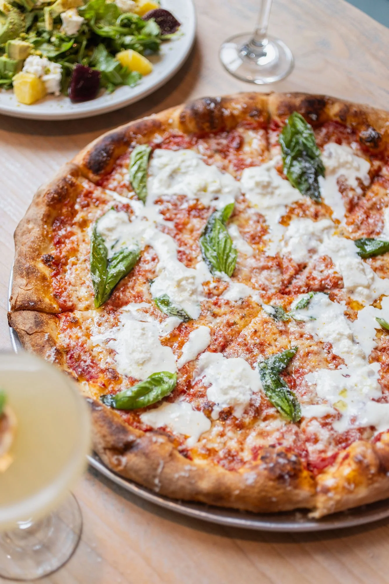 Close-up of a Margherita pizza with basil leaves, mozzarella cheese, and tomato sauce on a metal dish. In the background, there is a plate of salad with greens, cheese, and fruit pieces, and a glass of white wine. A partially visible glass of white wine is also in the foreground.