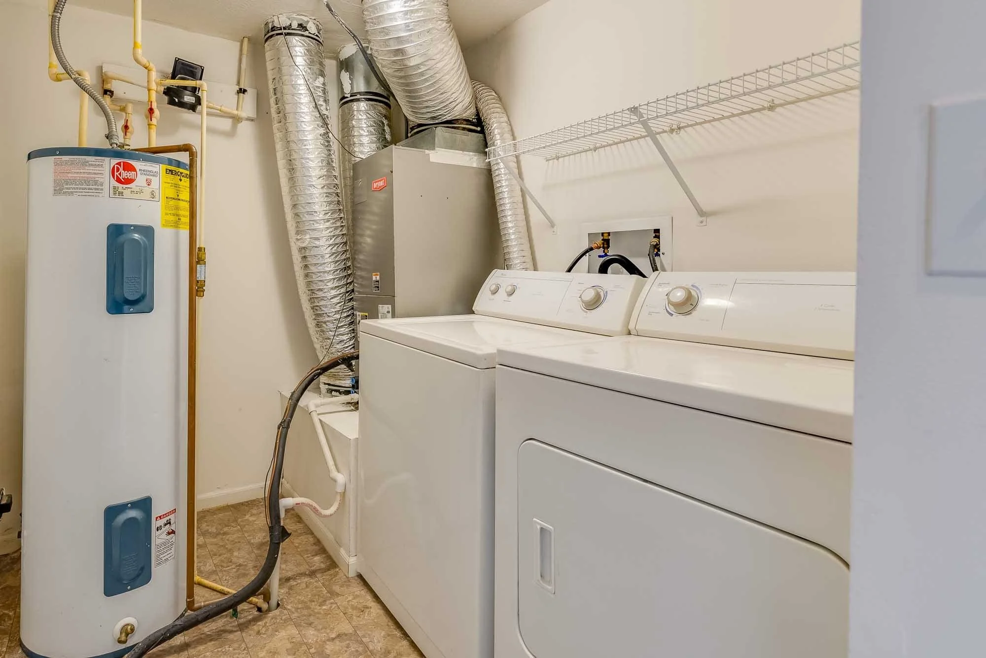 A laundry room containing a white water heater, a silver furnace with large flexible ductwork, a white washing machine, and a white dryer with control knobs. White wire shelving is mounted on the wall above the appliances.