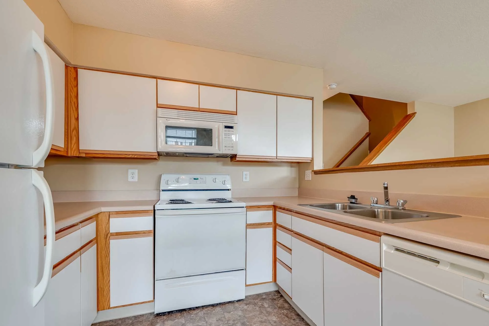 Kitchen with white cabinets, a white stove, a white microwave above the stove, a double sink with a faucet, and a beige wall. There is a partial view of a white refrigerator on the left and a double electrical outlet visible on the wall. The floor has a brown patterned tile. There is a staircase with wooden railings in the background.