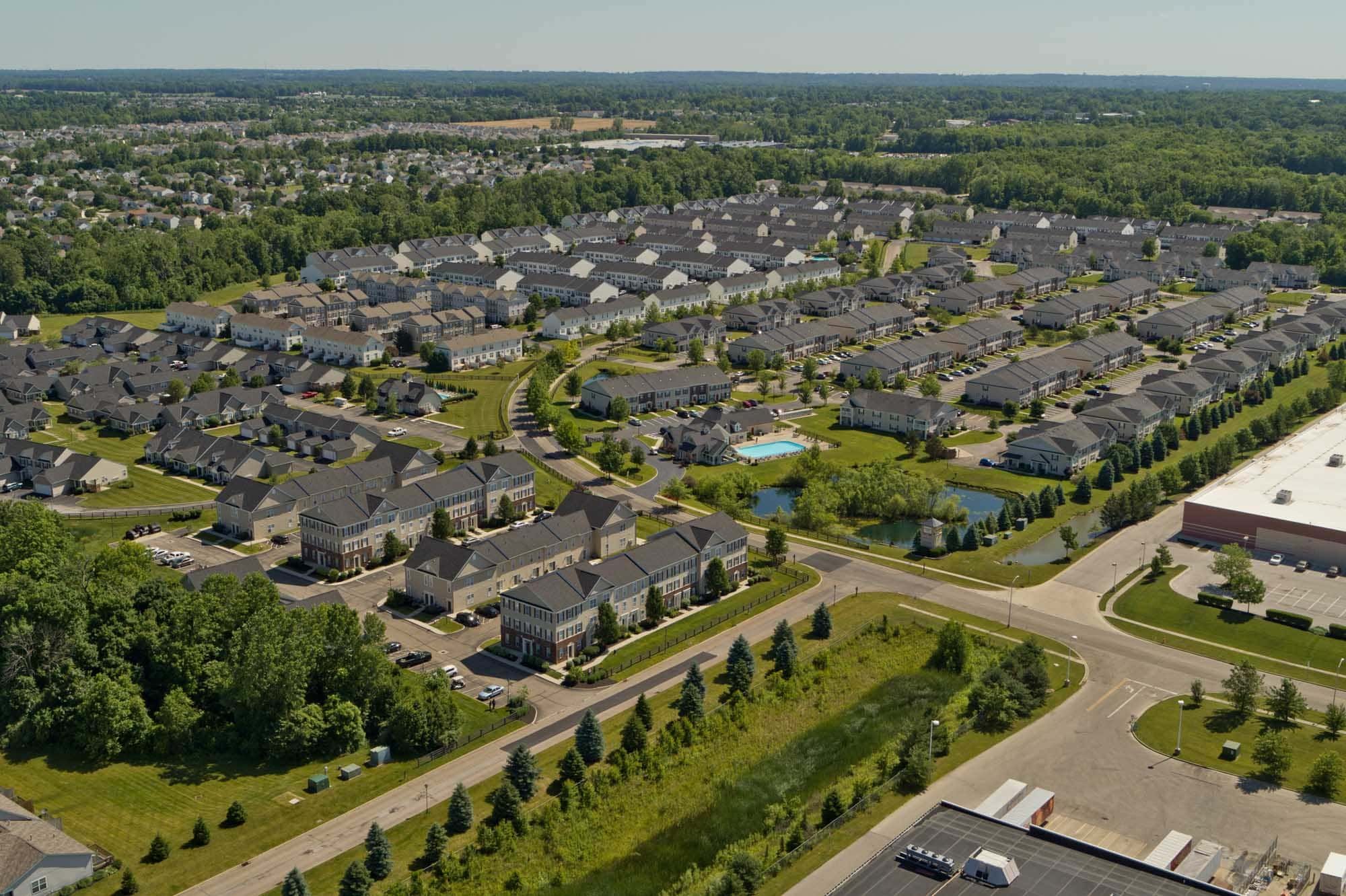 Aerial view of a suburban neighborhood with rows of townhouses, green lawns, and a swimming pool, surrounded by trees and a distant landscape.