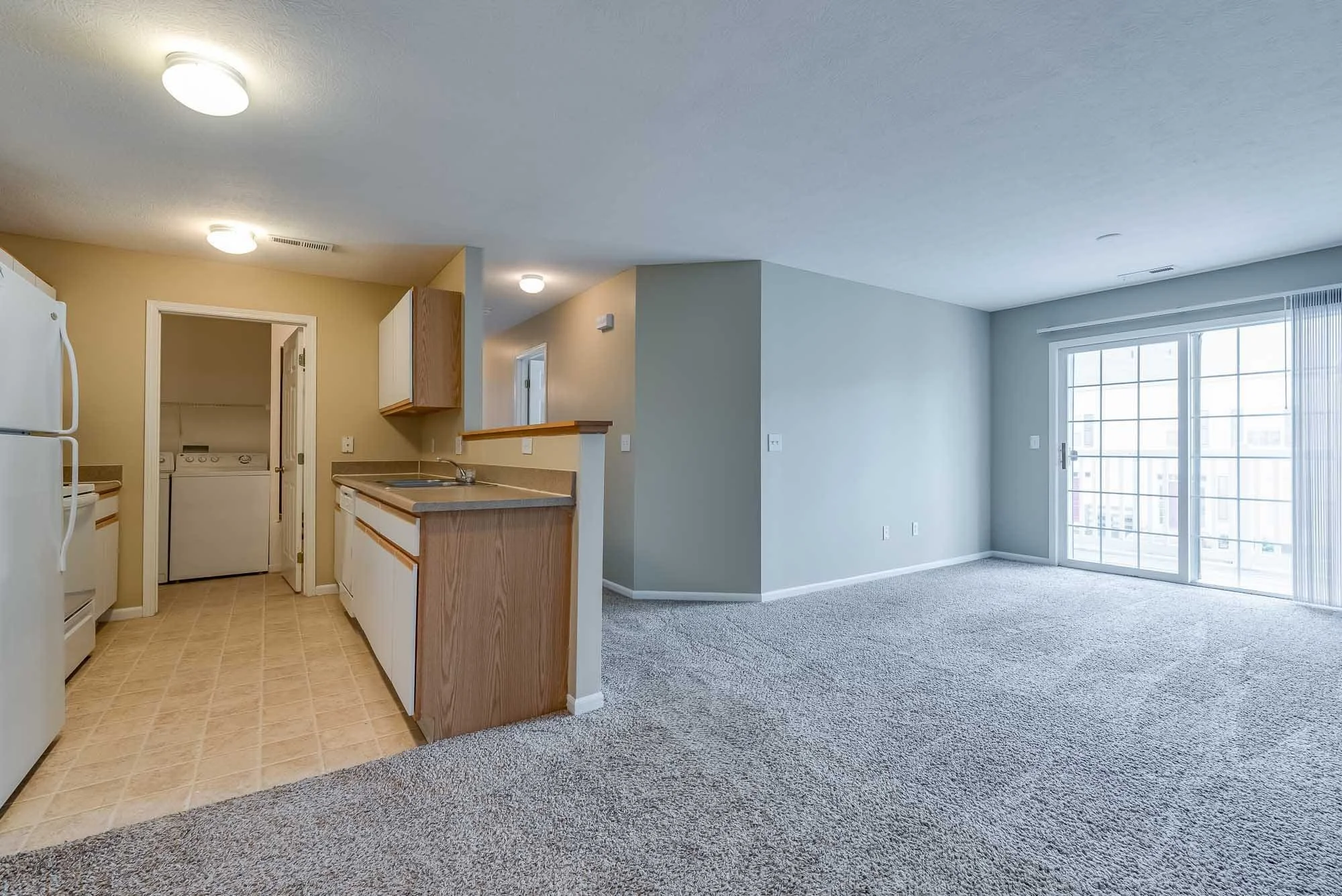 Interior of an apartment with a kitchen on the left and a living area with a large window and sliding glass door on the right. The kitchen has white appliances, beige cabinets, and counters, with a laundry area visible through an open door. The livin