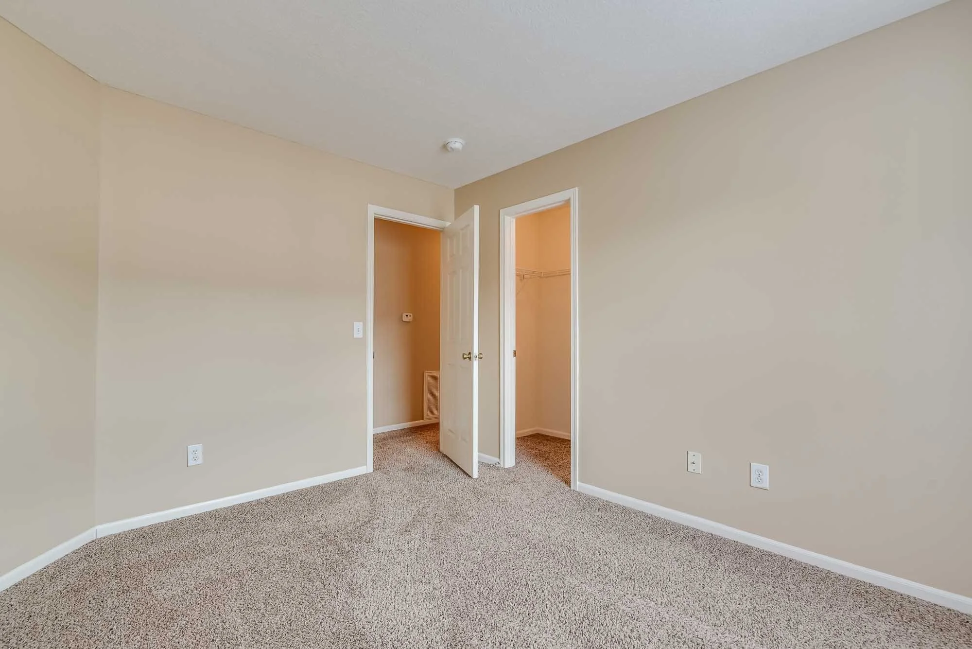 Empty beige bedroom with wall outlets, carpeted floor, open closet and door.