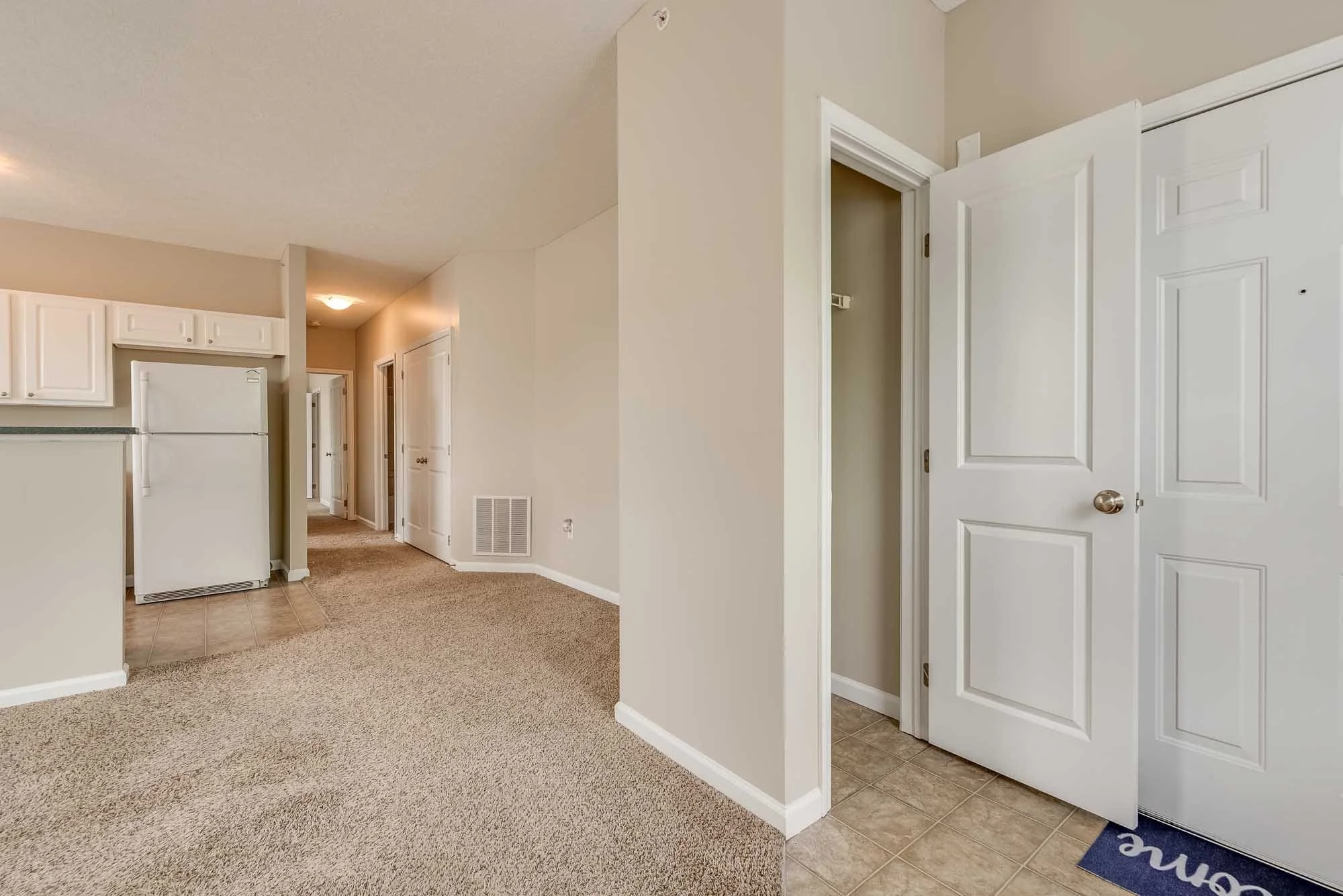 Interior of a modern apartment with open hallway, white kitchen cabinets, fridge, and a partly open door leading to another room.