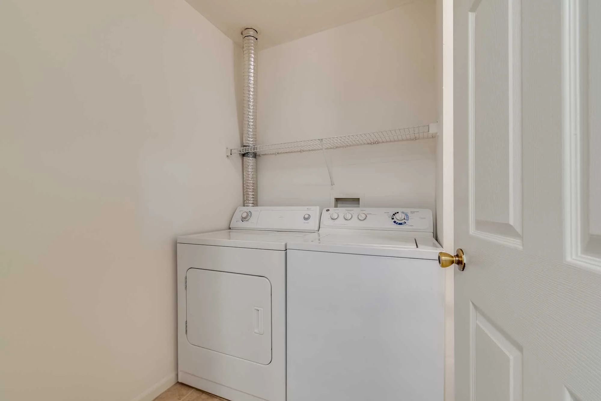 A laundry room with a white top-loading washing machine and a matching dryer. There is a wire shelf above the appliances and a flexible exhaust vent pipe running from the dryer to the ceiling. The room has a beige wall and a white door with a gold do