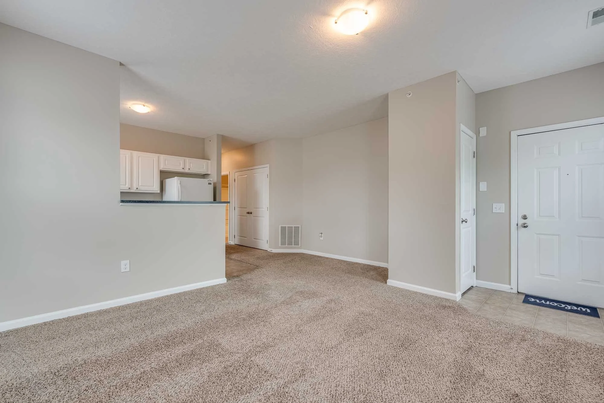 Interior view of a living area with beige carpet, white walls, and a white front door, with a partial view of the kitchen with white cabinets and appliances.