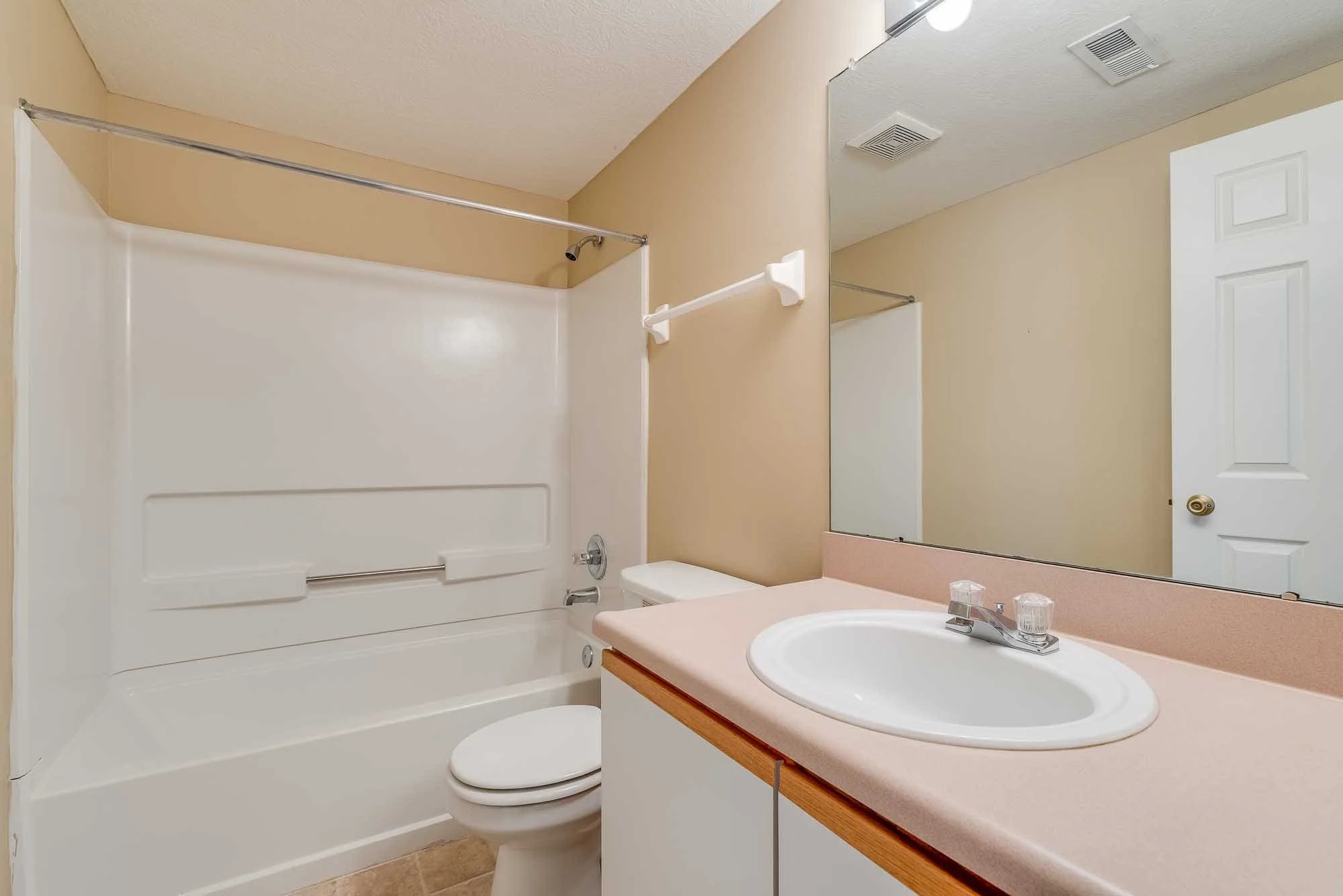 A small bathroom with a bathtub, toilet, and sink. The walls are painted beige, and there is a large mirror above the sink. The ceiling has a vent and recessed lighting. The countertop is pink, and the sink has a single faucet.