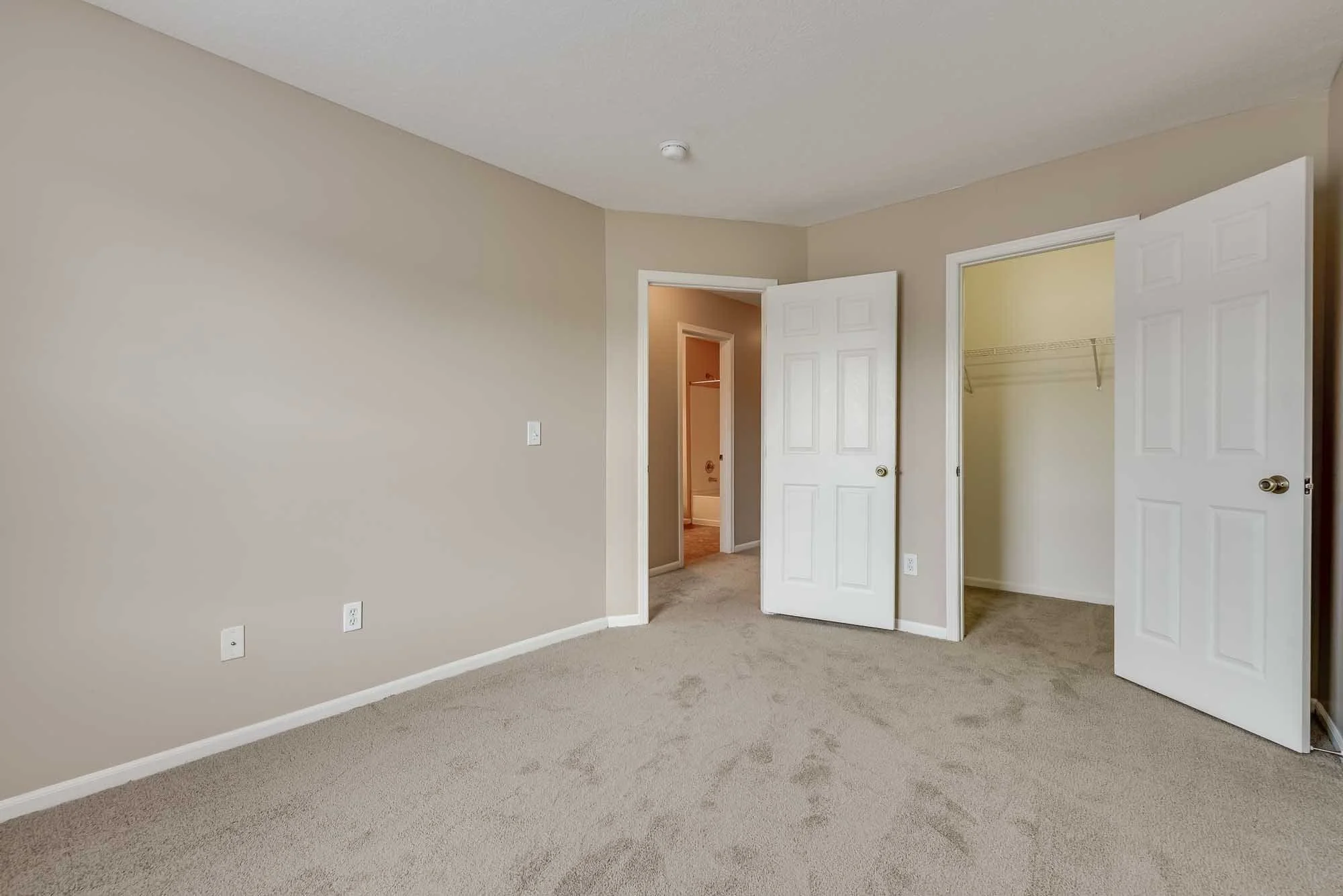 Empty bedroom with beige walls, beige carpet, and white trim. Two open doors reveal walk-in closets, and a door leading to a hallway or bathroom.