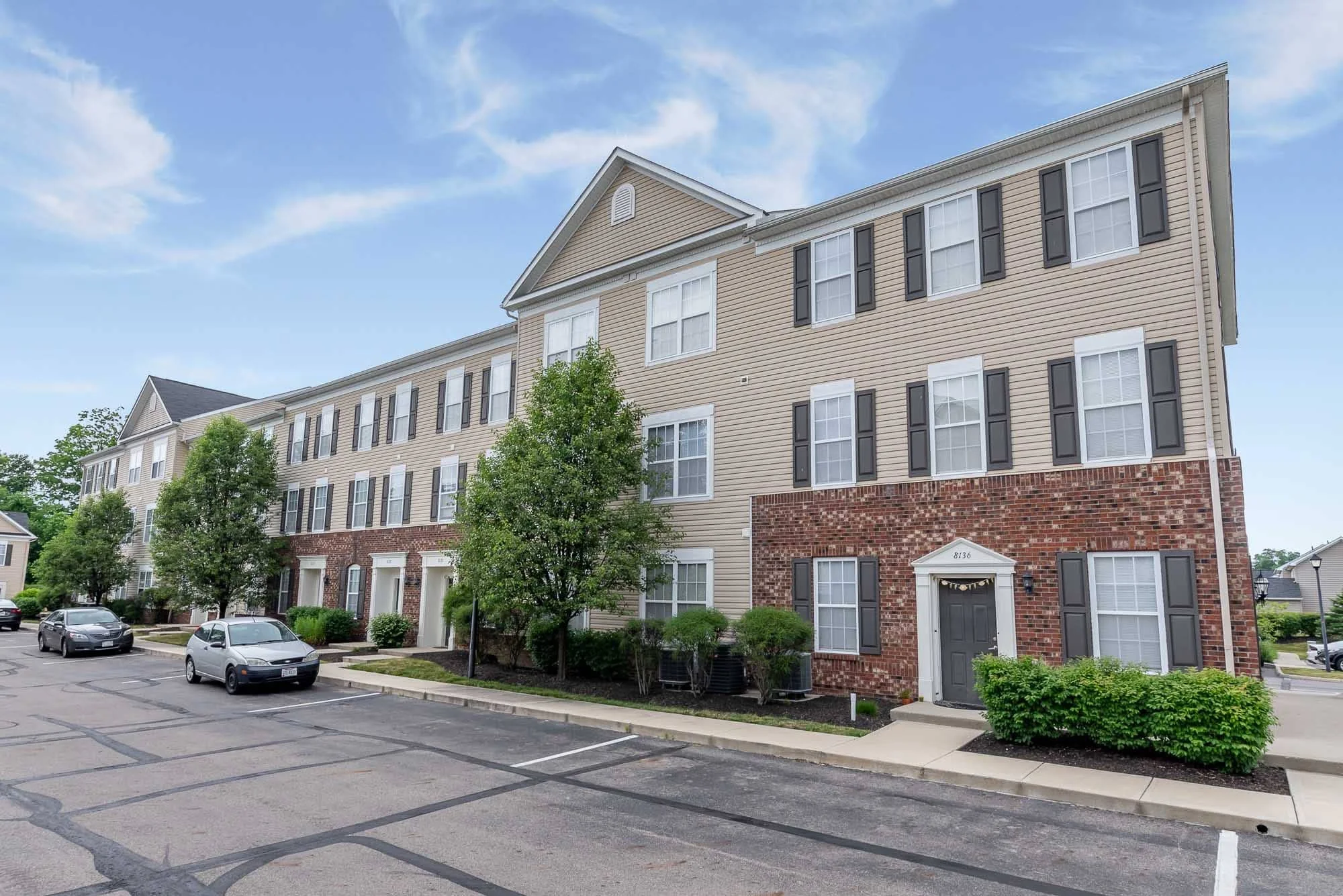 Exterior view of a multi-story apartment building with beige siding, black window shutters, and a brick foundation, situated near a parking lot with parked cars, green trees, and a partly cloudy sky.