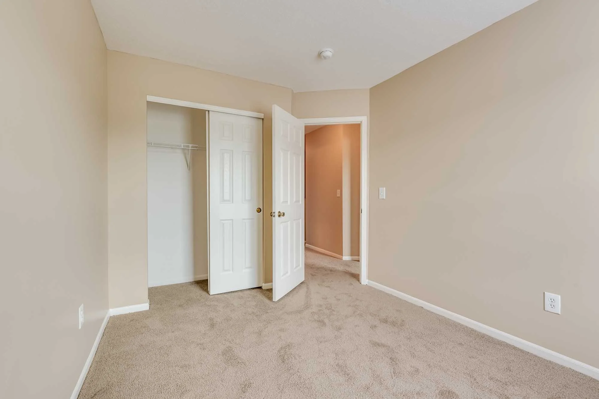 Empty bedroom with beige walls and light brown carpet, open closet door, and doorway to hallway.