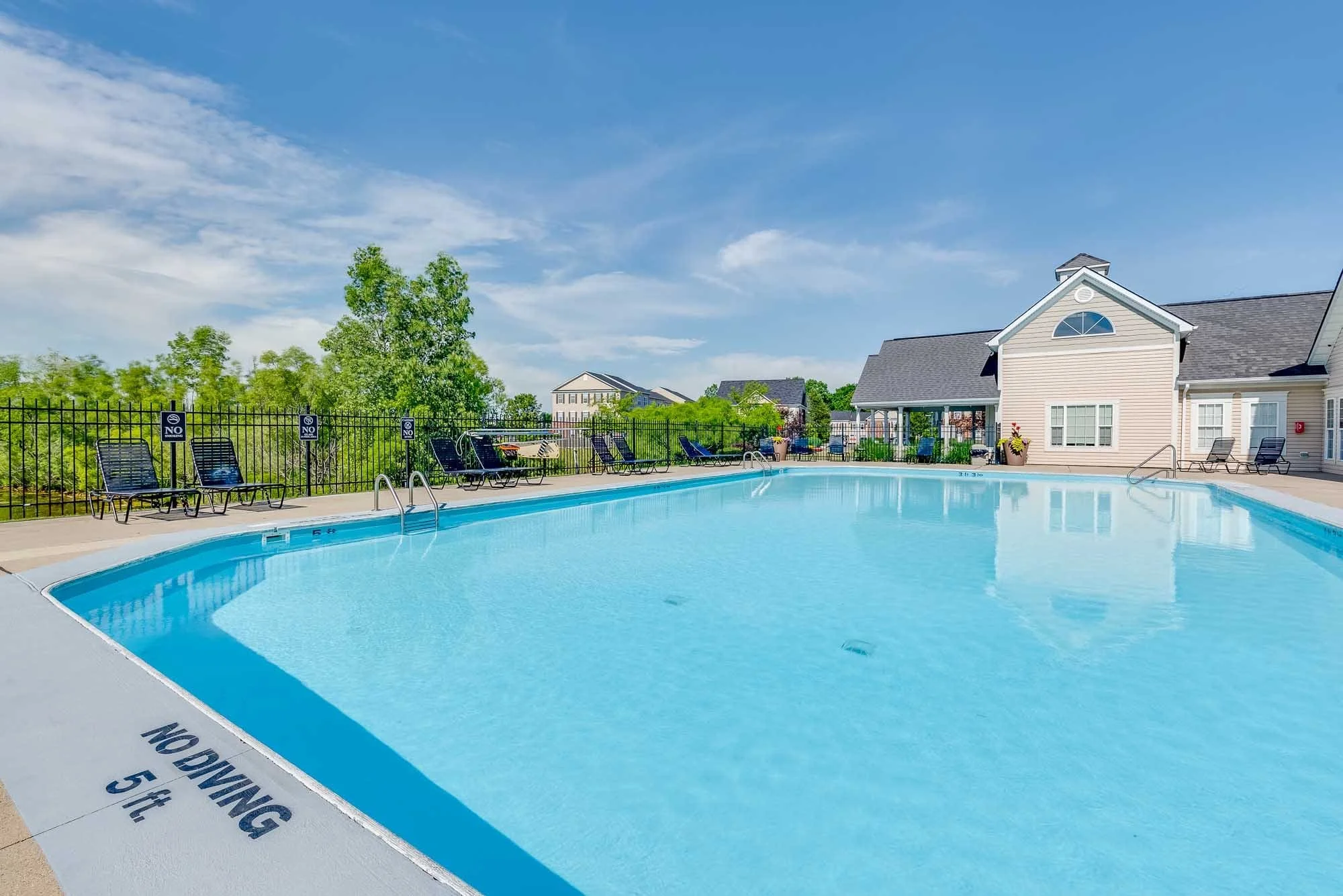 Community swimming pool with lounge chairs along the side, surrounded by greenery and a black fence, under a blue sky with some clouds, in a residential area with houses in the background.