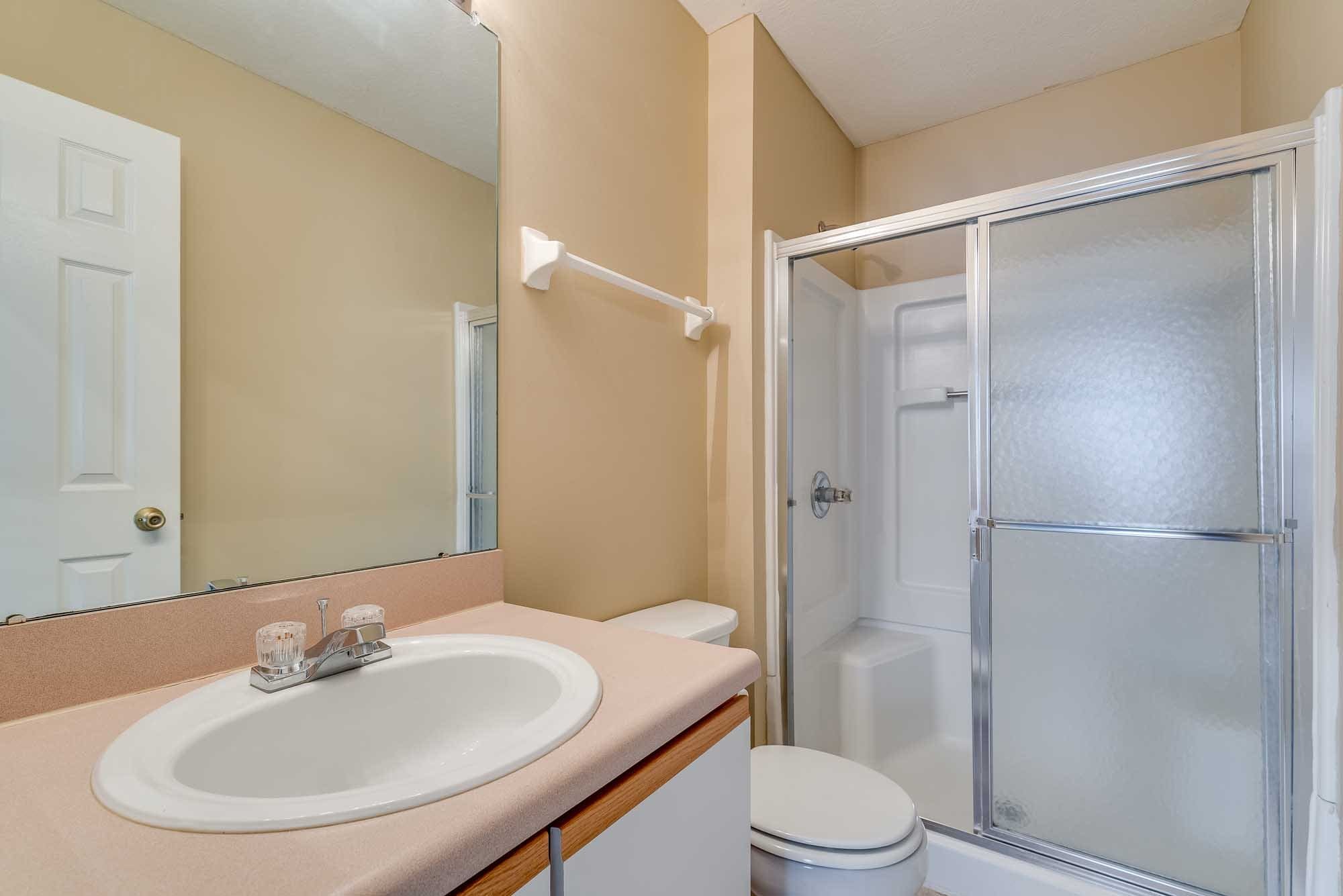 A bathroom featuring a sink with a beige countertop, a large mirror, a white shower stall with frosted glass doors, and a beige wall with a white towel rack.