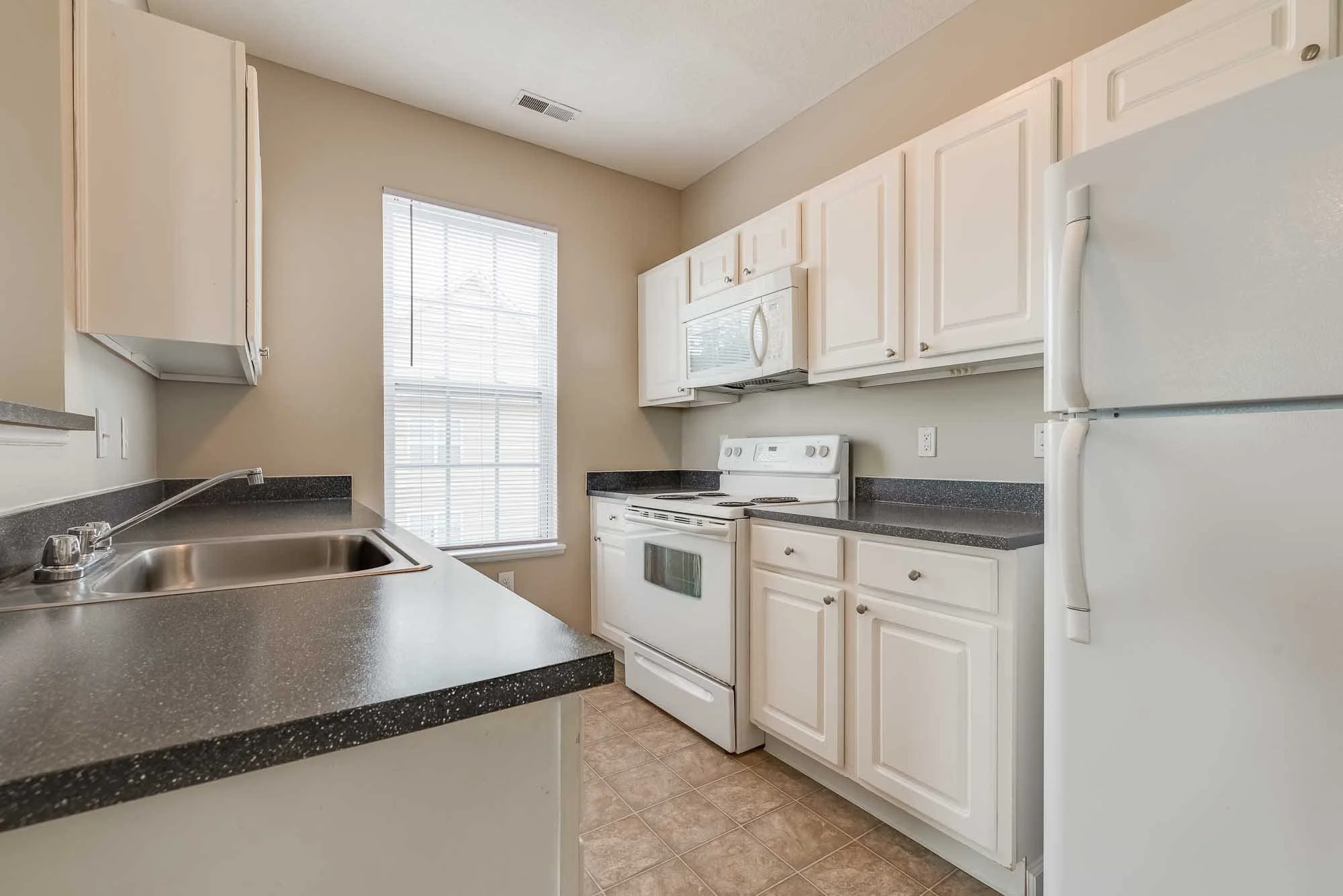 Kitchen with white cabinets, black speckled countertops, white appliances including a refrigerator, oven, and microwave, a stainless steel sink, beige walls, a window with blinds, and tile flooring.