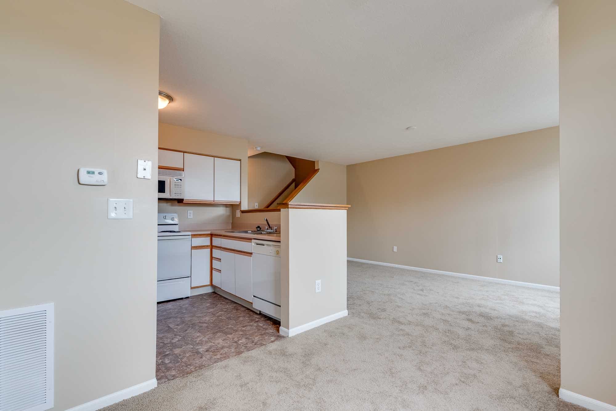 An open floor plan living space with beige walls, a kitchen area with white appliances and light wood cabinets, and a carpeted living area. There is a staircase with a wooden railing leading to an upper level.