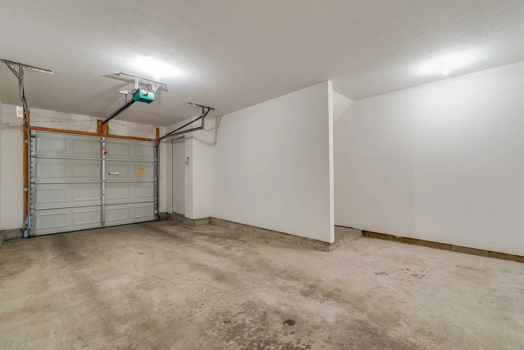 Empty garage with concrete floor, white walls, and a white garage door, with drywall dividers and garage door mechanisms visible.
