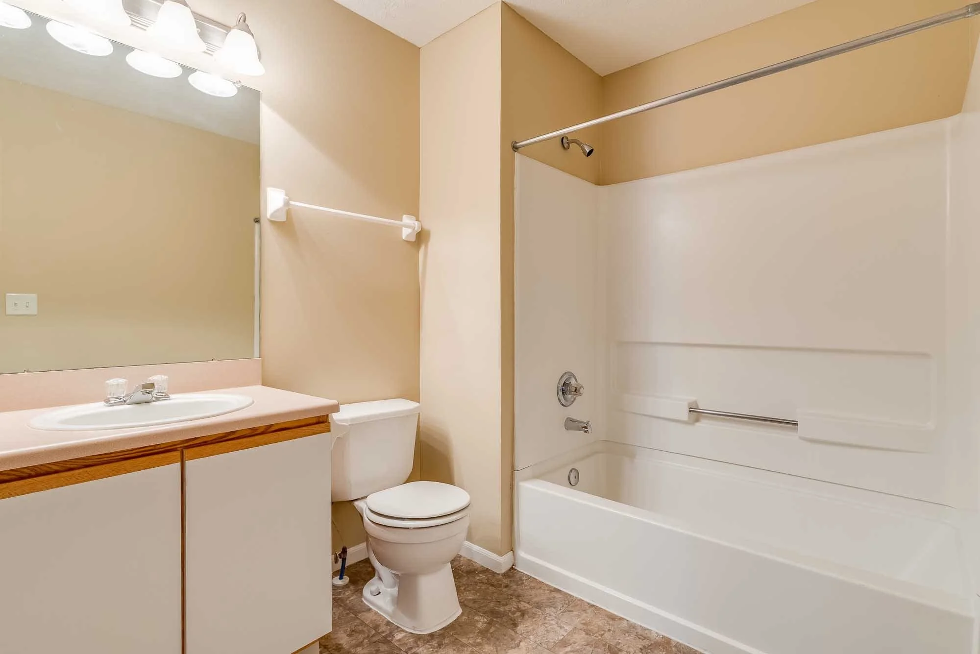 A bathroom with a mirror, light fixtures, a sink with a wooden cabinet, a toilet, and a bathtub with a shower curtain rod.