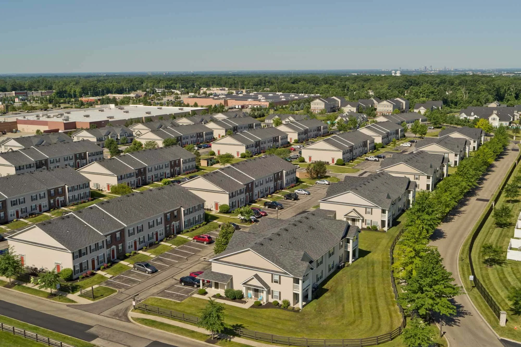 Aerial view of a residential apartment complex with multi-story townhouses, parking lots filled with cars, surrounded by green trees and a well-maintained lawn, with a city skyline in the distance and clear blue sky.