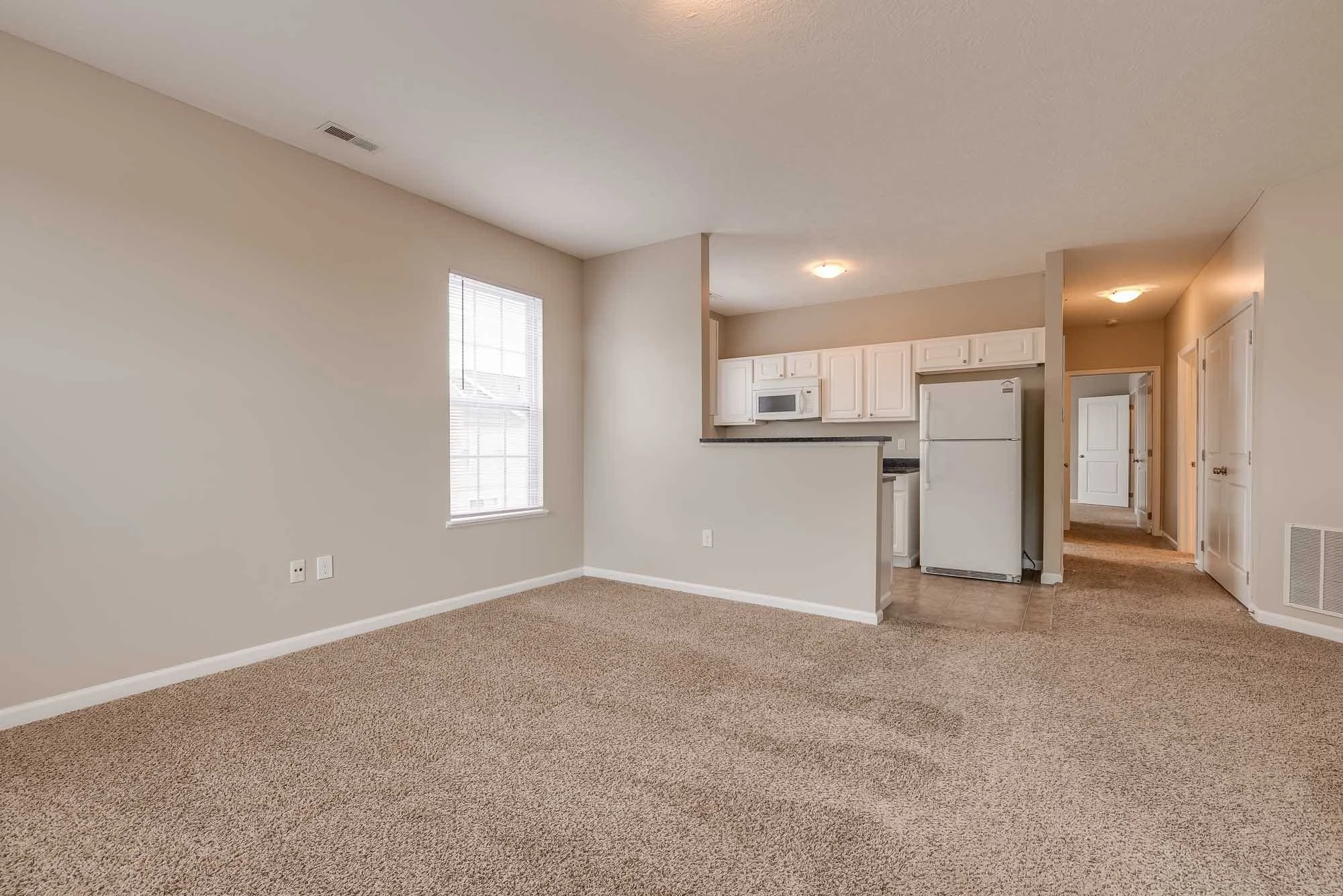 Interior of an unfurnished apartment featuring an open living area with beige carpeting and walls, and a kitchen with white cabinets, a microwave, and a refrigerator.