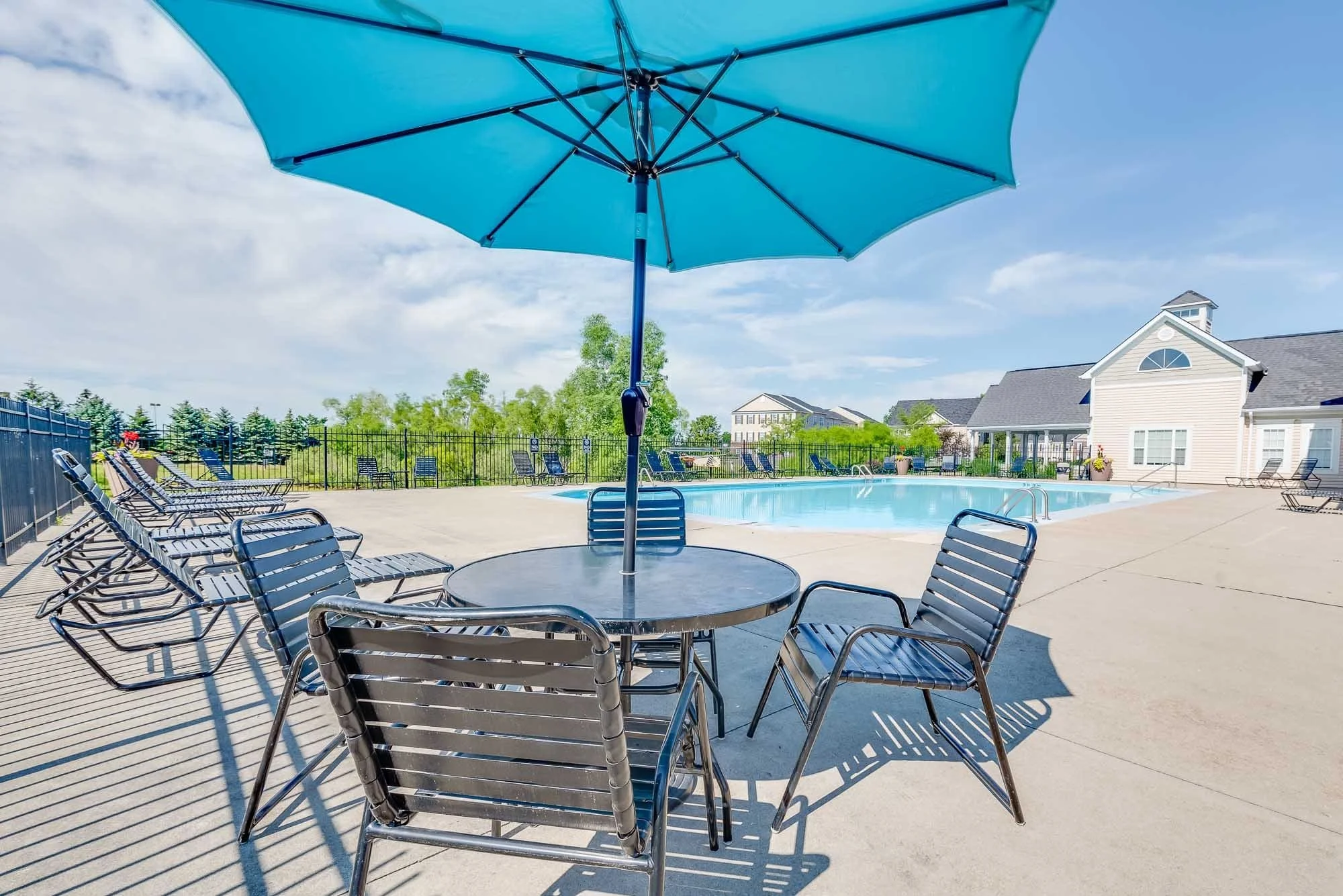 A swimming pool surrounded by a concrete deck with lounge chairs and a large blue patio umbrella in the foreground. In the background, there are buildings, trees, and a clear blue sky with some clouds.