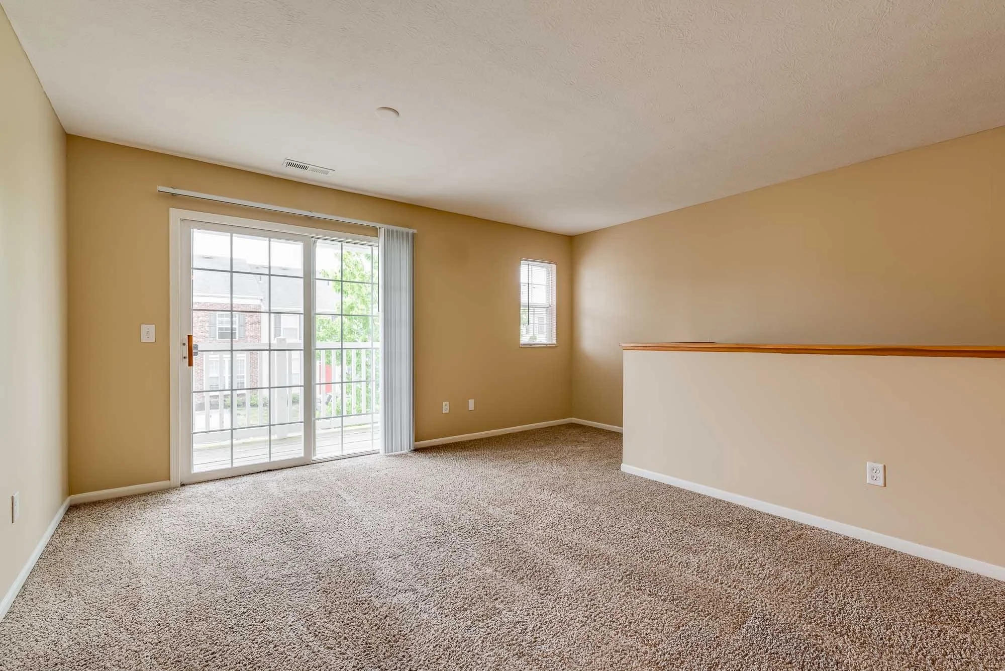 Interior view of an empty living room with beige walls, carpeted floor, sliding glass door leading to a balcony, and two small windows, with sunlight coming in.