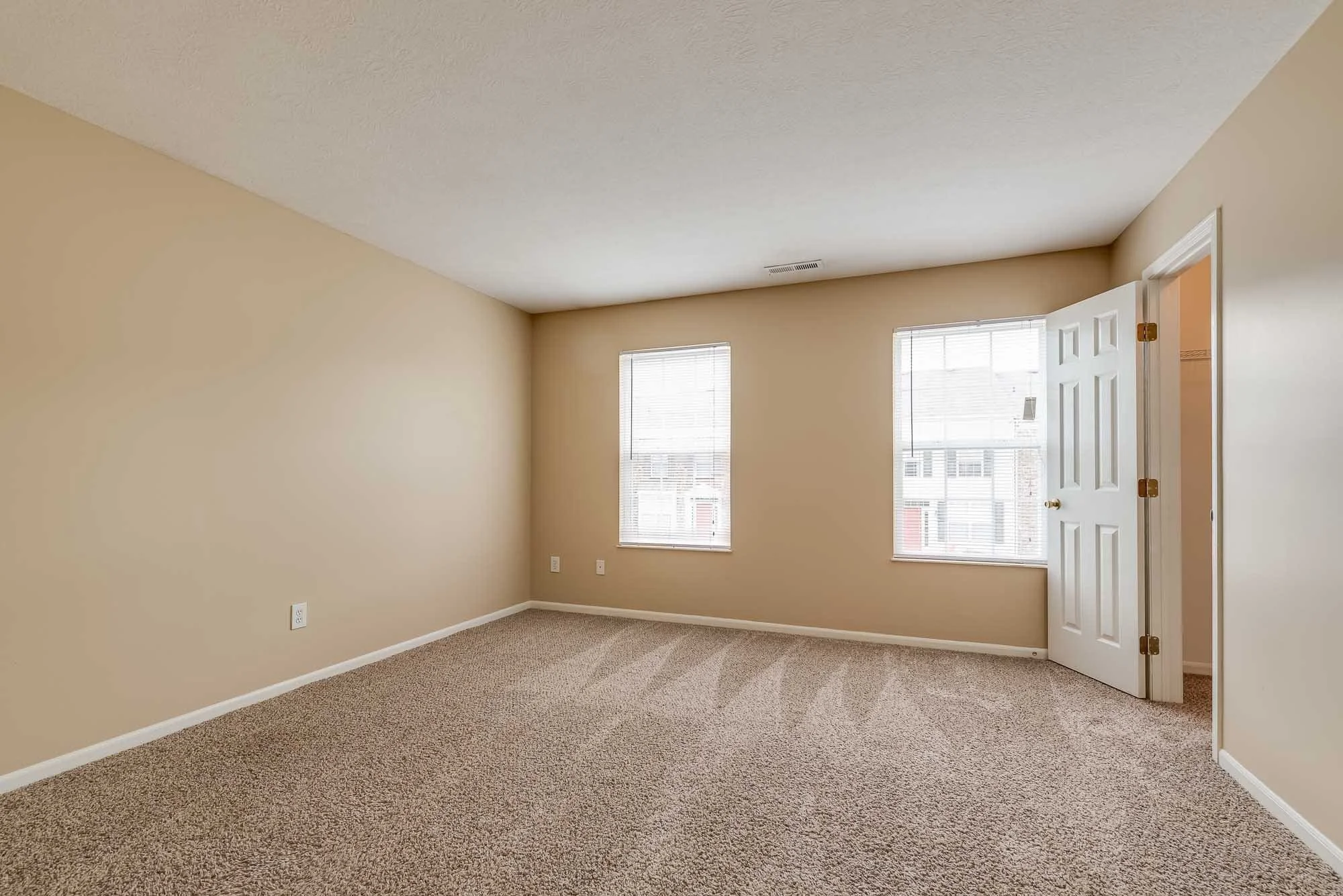 Empty beige-colored room with two windows, an open door, and light-colored carpeting.