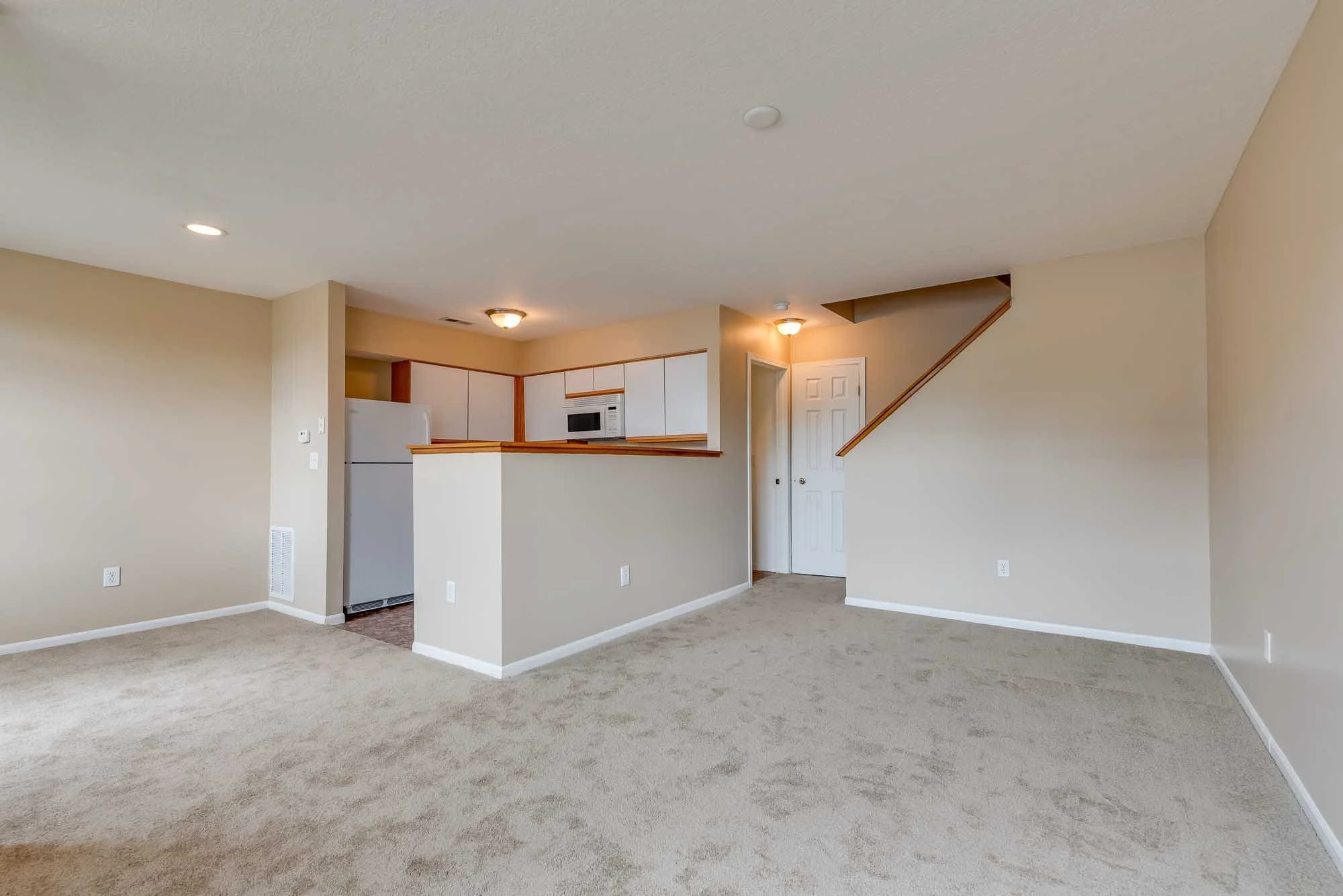 Interior living room with beige walls, tan carpet, open kitchen with white cabinets, white appliances, and a doorway leading to another room.