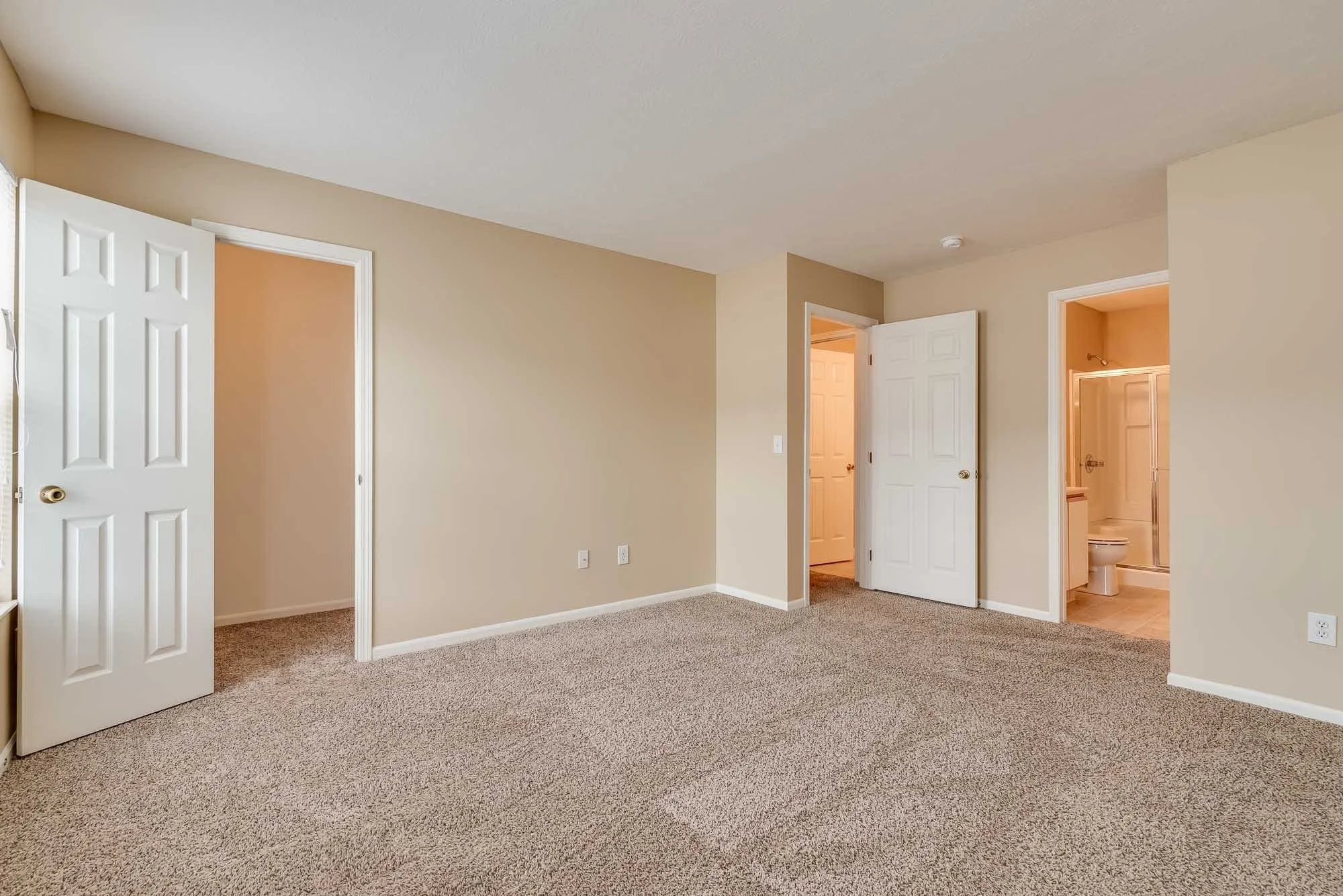 Empty beige carpeted room with multiple open white doors leading to bedrooms and bathroom.