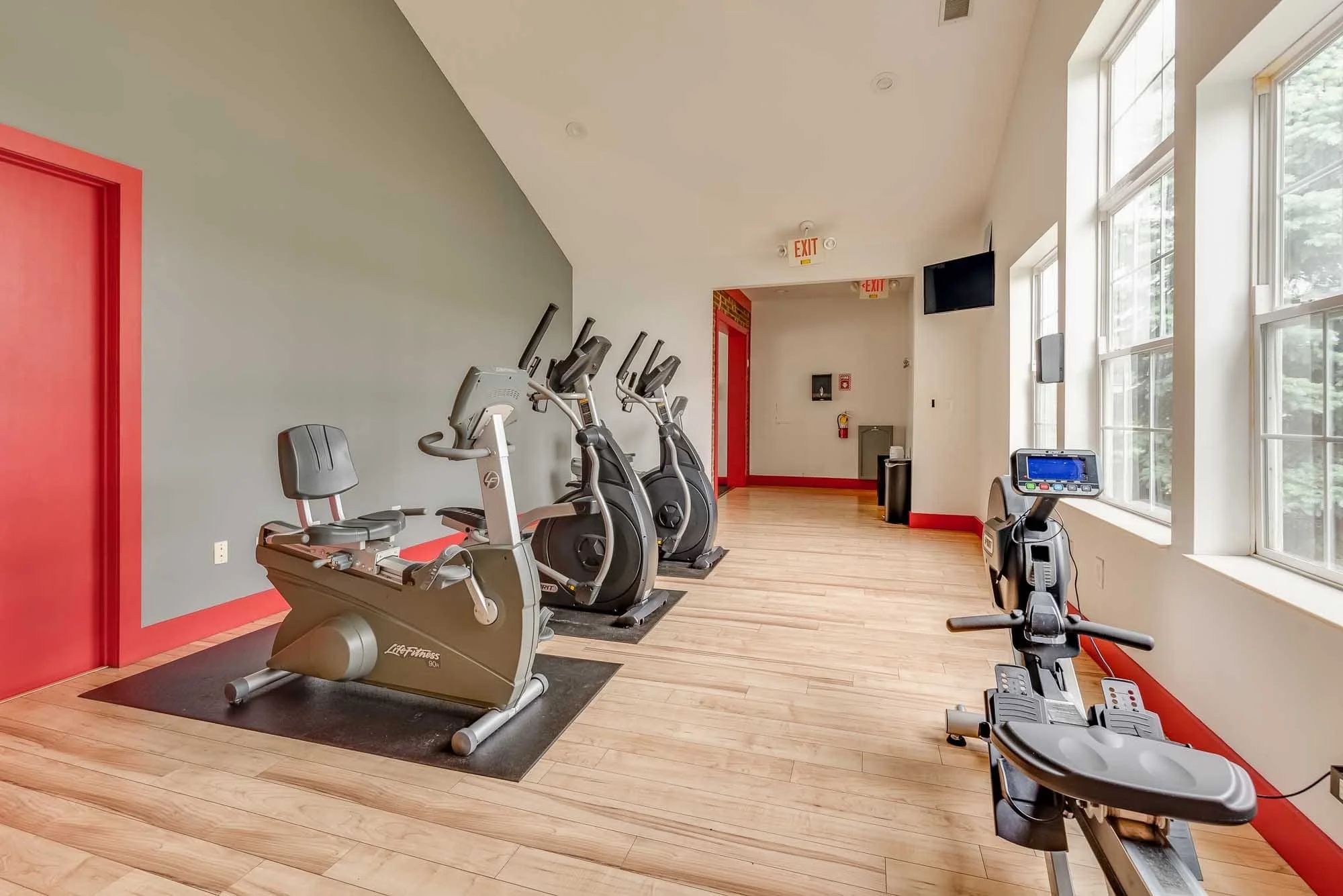 A gym with cardio equipment including ellipticals and stationary bikes, large windows, a TV mounted on the wall, and wooden floors.