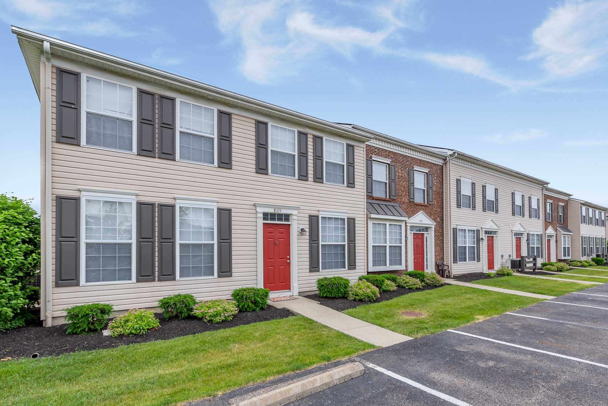 Row of beige townhouses with red front doors, gray shutters, and small landscaped gardens, under a partly cloudy sky.