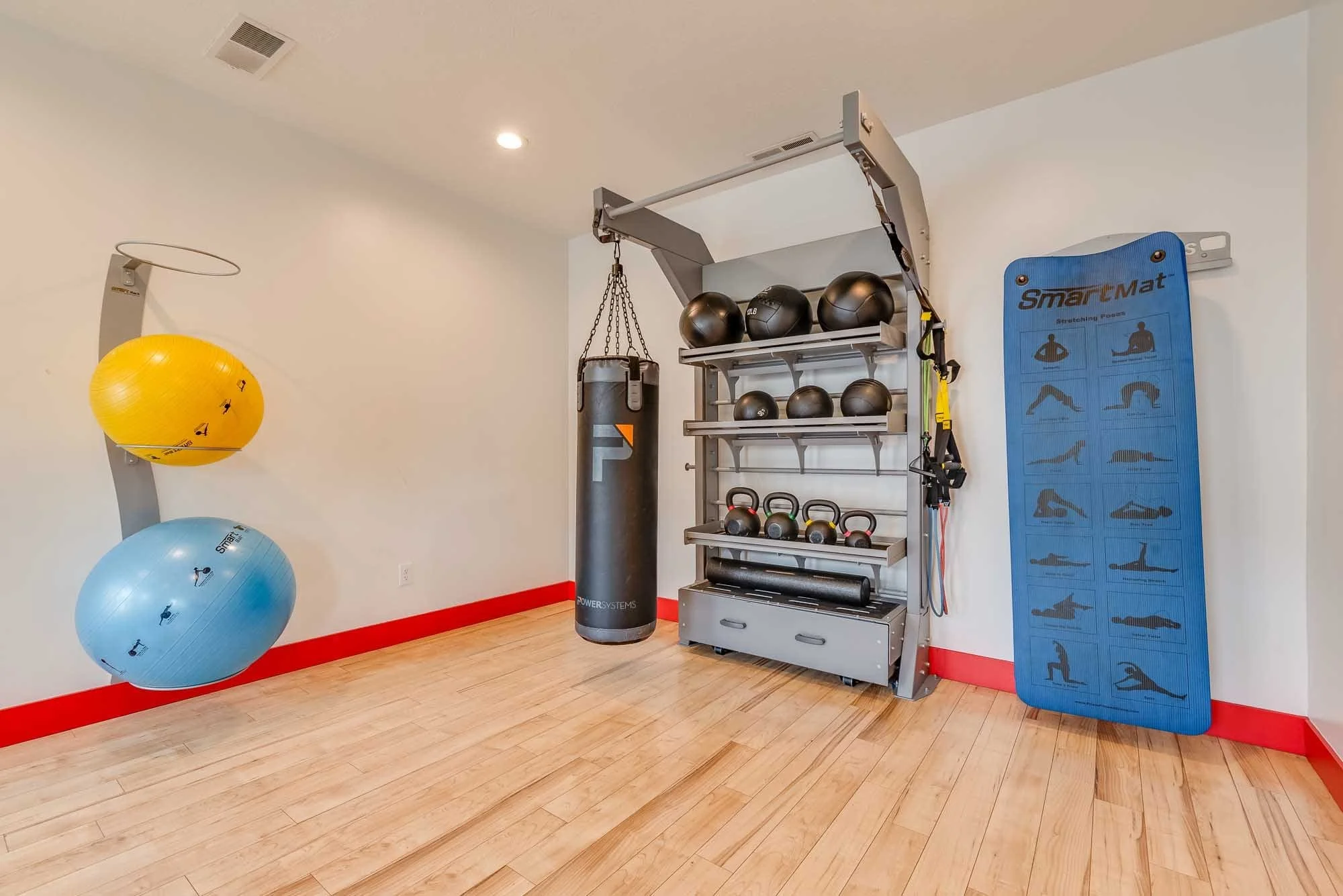 Fitness center with exercise balls, a punching bag, kettlebells, resistance bands, foam rollers, and an exercise mat. The room has white walls with red trim and a hardwood floor.