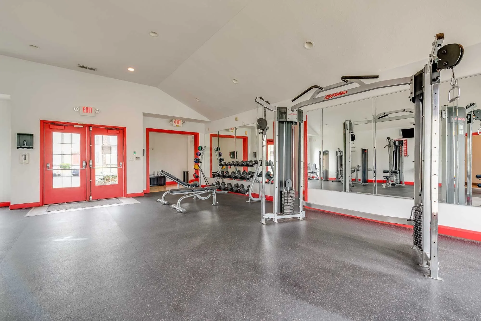Interior of a modern gym with red doors, mirrors, and various workout equipment including dumbbells and machines.