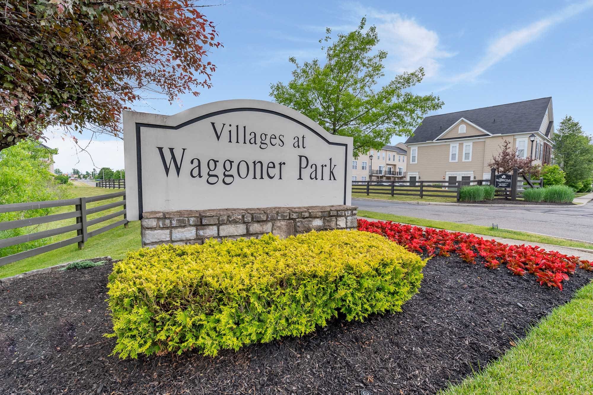 Sign at Villages at Waggoner Park surrounded by colorful flowers and bushes with buildings and trees in the background.
