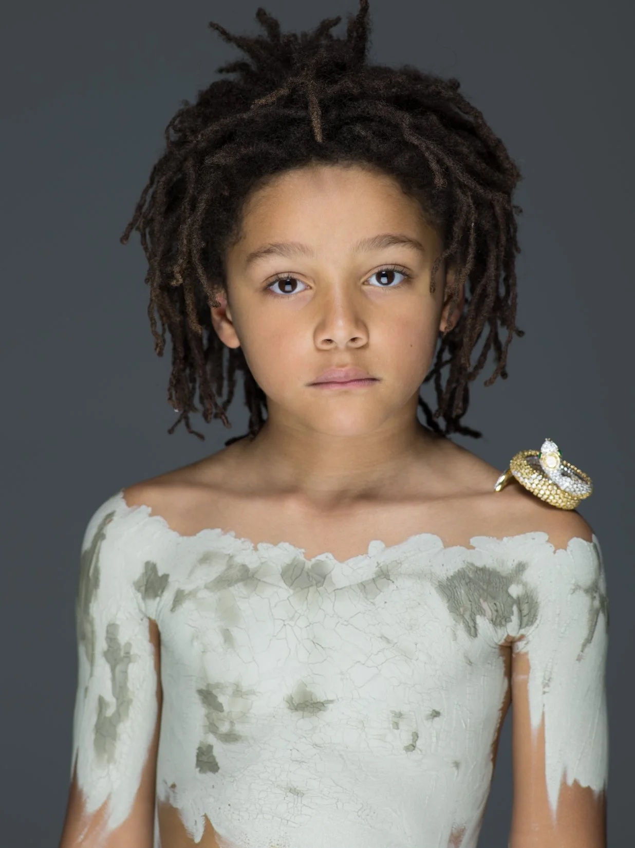 Ragazza con capelli ricci e dreads, vestita con un abito bianco sfilacciato e graffiato, indossa un anello decorativo sulla spalla, sfondo grigio scuro.
