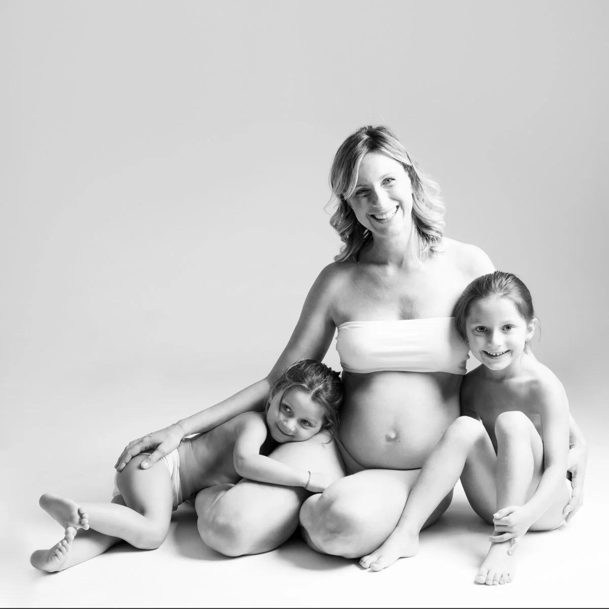 Una donna incinta con due bambine in posa su uno sfondo bianco, sorridenti e abbracciate, in una foto in bianco e nero.